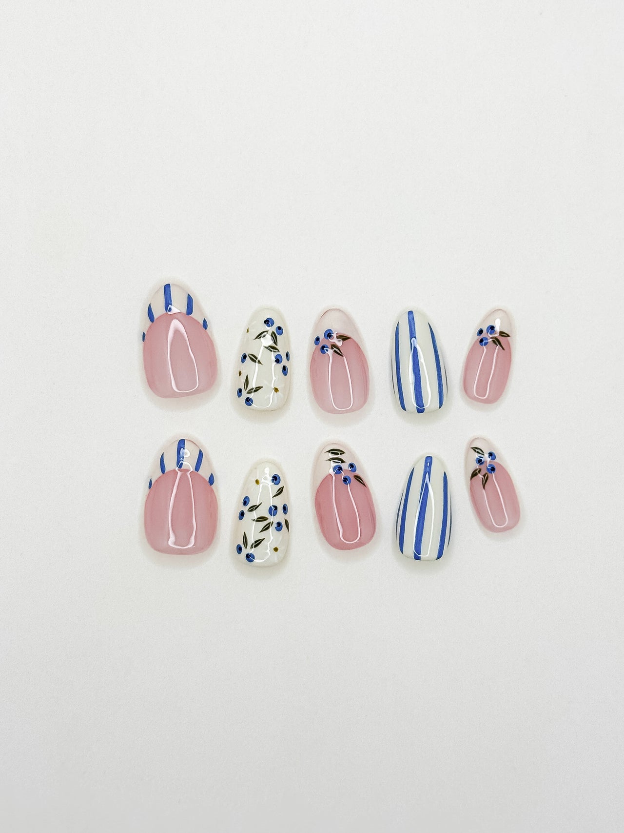 Blue and white Mediterranean-inspired press-on nails with hand-painted floral detailing and striped accents in a short almond shape. Handmade reusable false nails inspired by European summer nails, coastal Italian nail art and vintage ceramic patterns.