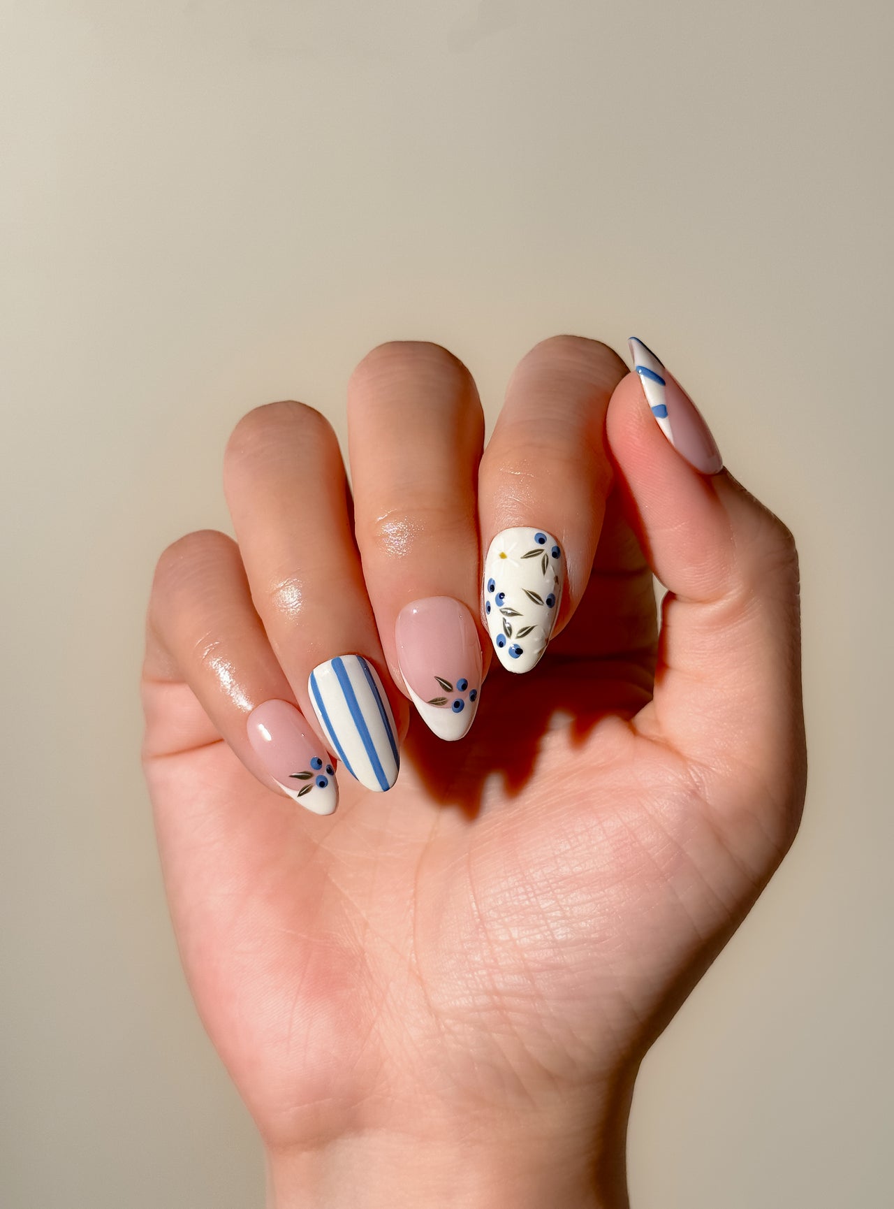 Blue and white Mediterranean-inspired press-on nails with hand-painted floral detailing and striped accents in a short almond shape. Handmade reusable false nails inspired by European summer nails, coastal Italian nail art and vintage ceramic patterns.