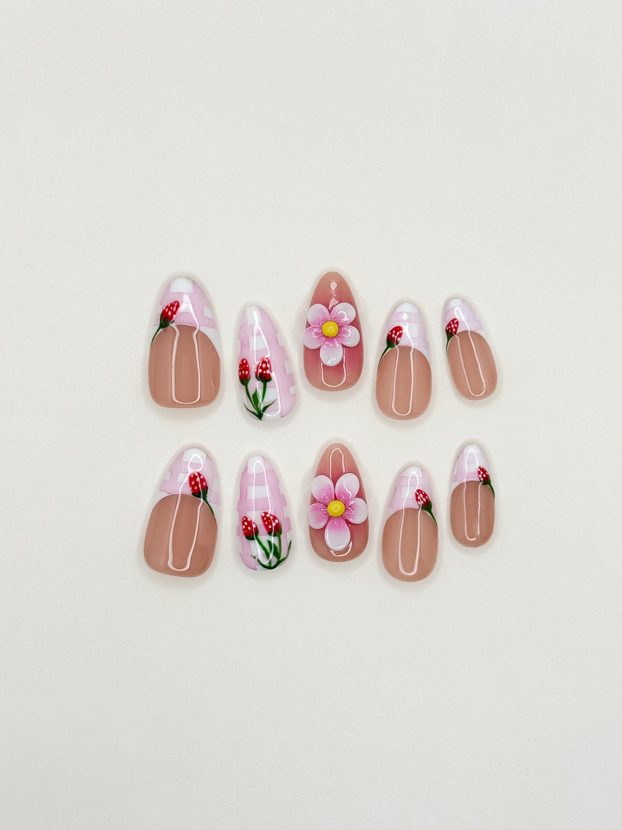 Strawberry Dream handmade reusable press-on nails laid flat, featuring sheer nude bases, pink gingham French tips, hand-painted strawberry details and raised 3D flower accents in a medium almond shape.