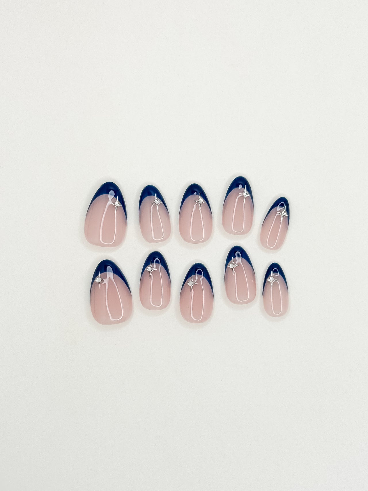 After Midnight handmade press-on nails arranged on a white background, featuring short almond nails with deep navy skinny French tips, a sheer blush base and silver star embellishments.
