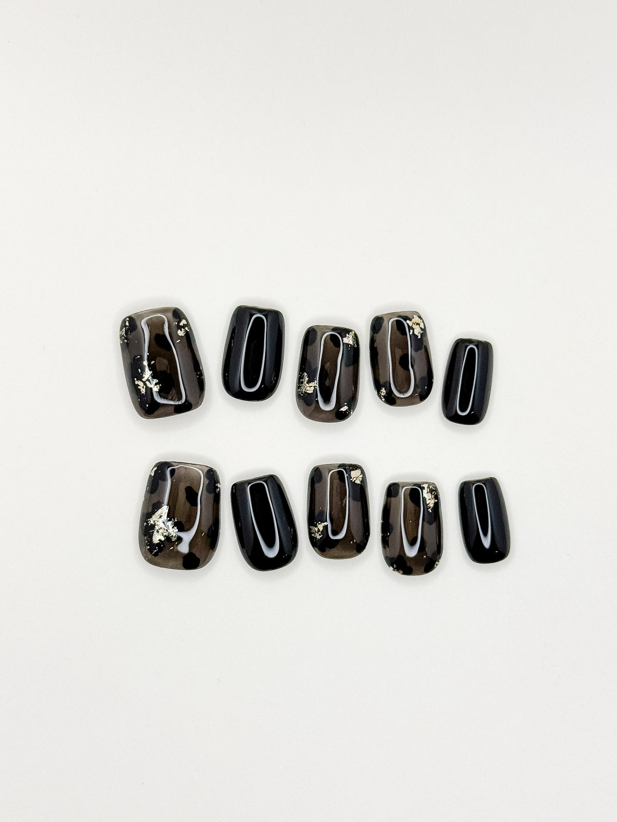 Allegedly smoky black short square press-on nails with translucent abstract panels and gold foil arranged on white.