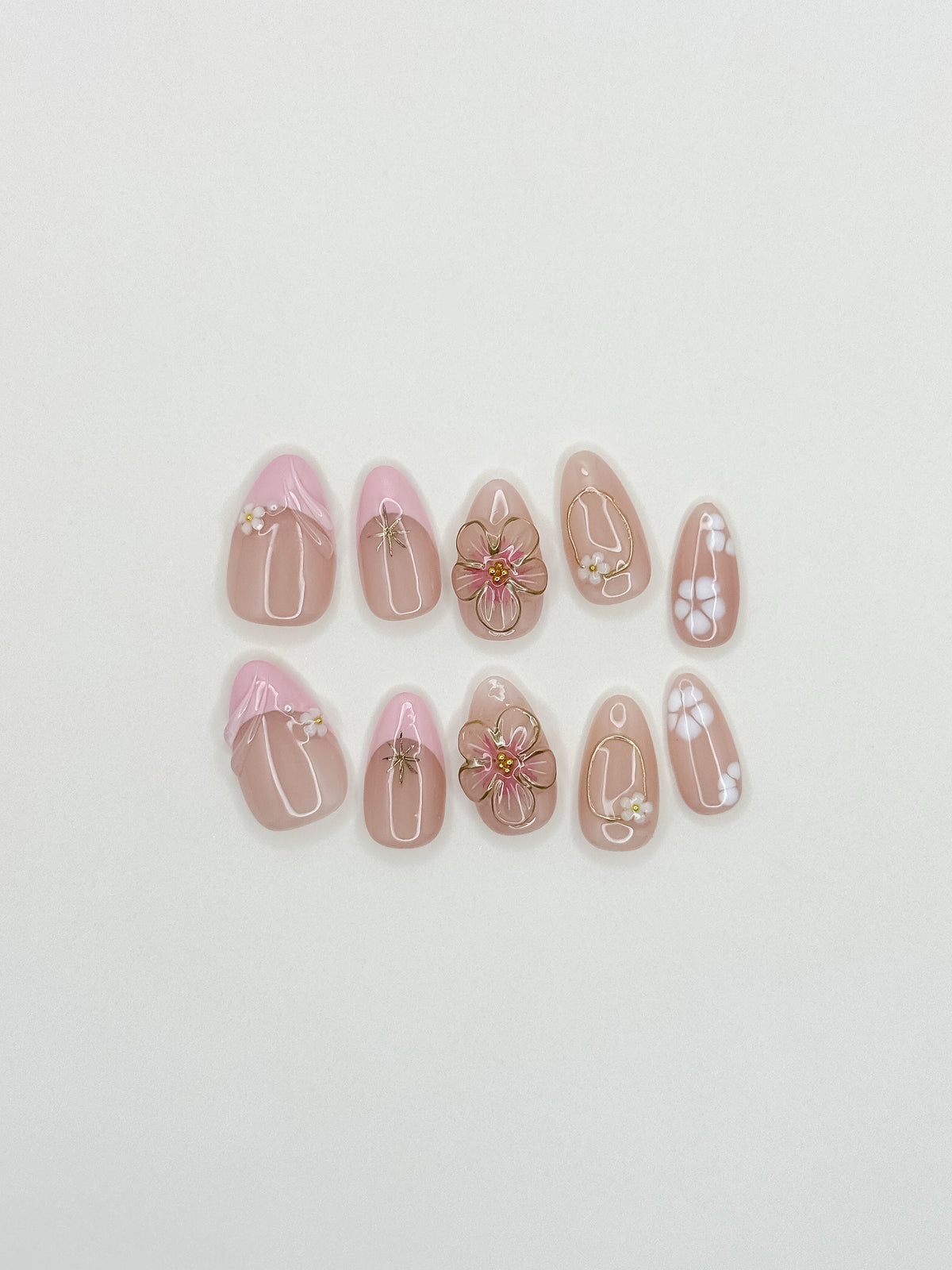 Petal handmade reusable press-on nails laid flat, featuring blush-pink bases, raised 3D flowers, white petals and gold details in a short almond shape.