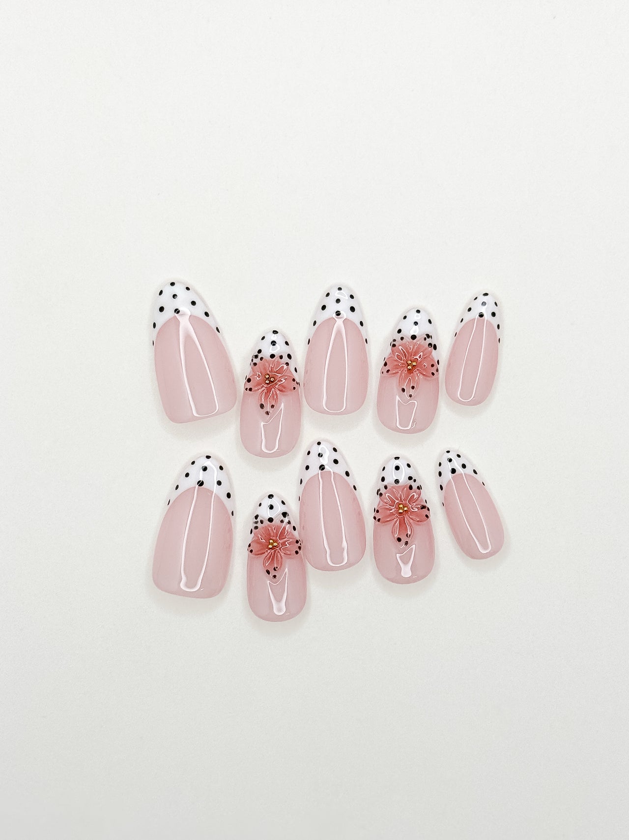 A set of ten false press-on nails arranged on a plain white background. The nails feature a glossy pale pink base with white curved stripes, black polka dots along the tips, and a small pink floral accent on two of the nails.