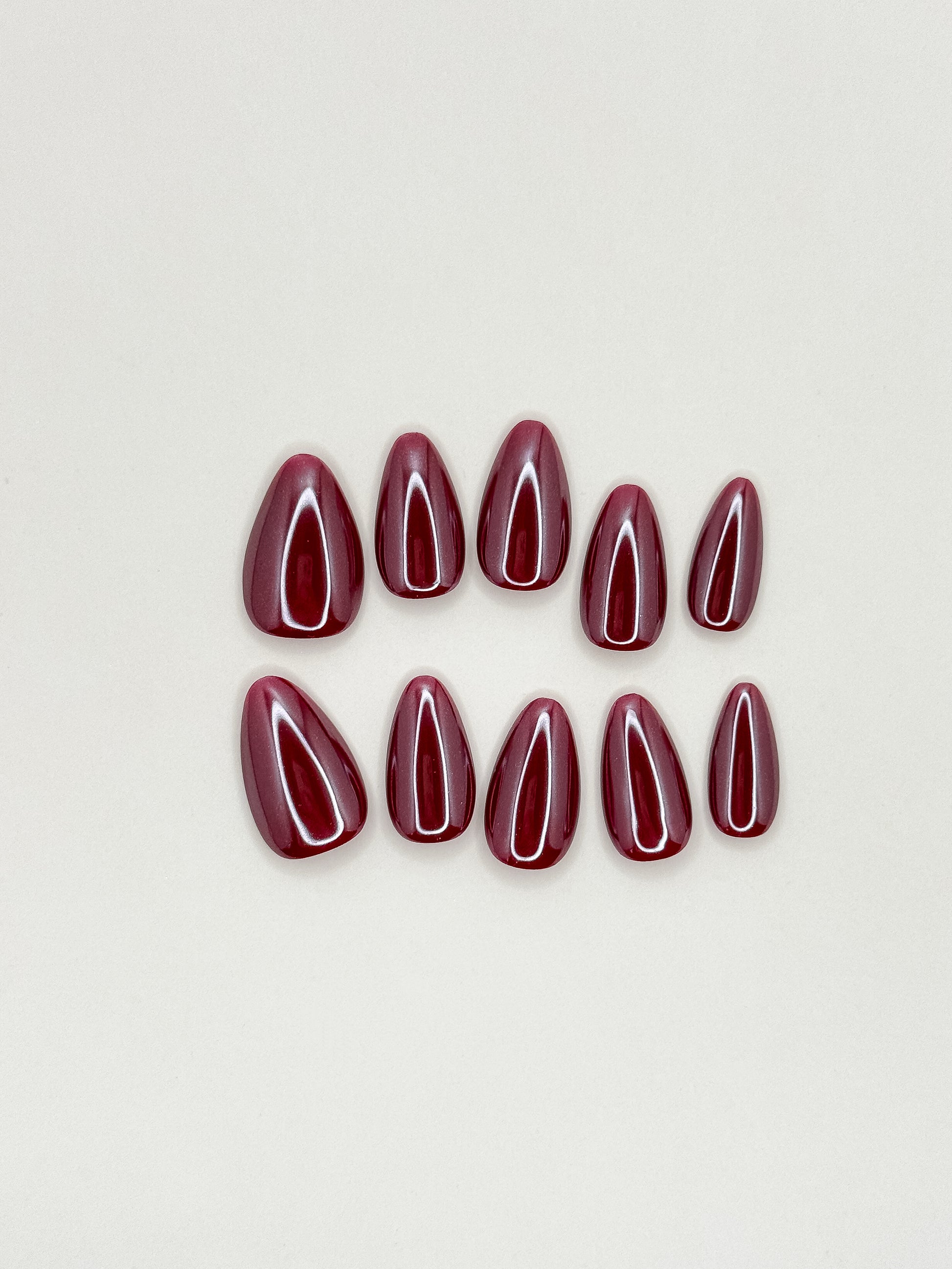 Deep berry red glazed press-on nails in a glossy short almond shape with reflective blackcurrant tones. Handmade reusable false nails inspired by cherry glaze nails, dark red manicures and glossy wine-coloured nail trends.