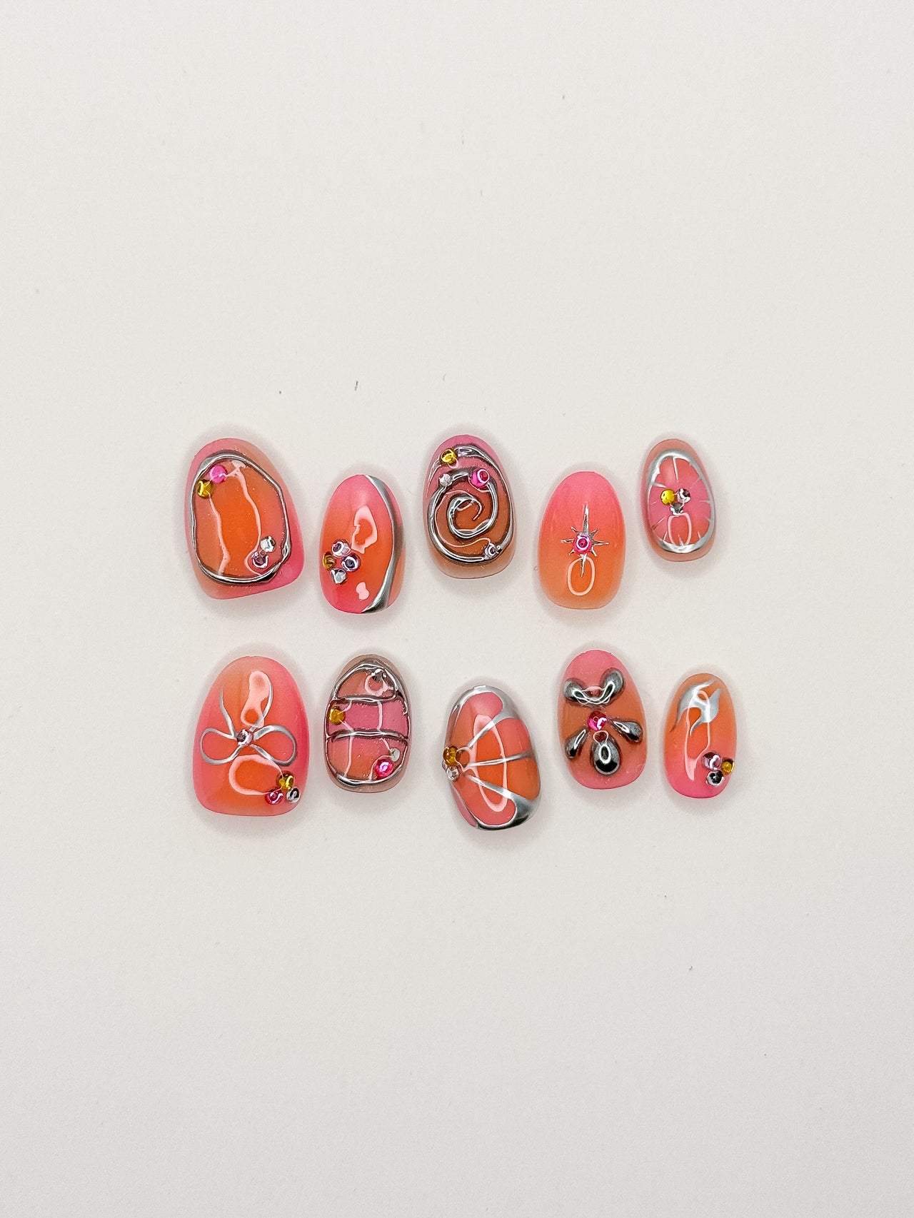 Coral Crush handmade reusable press-on nails laid flat, featuring coral and pink ombré nail art, chrome outlines, abstract swirls, floral motifs and gem accents in a extra short oval shape.