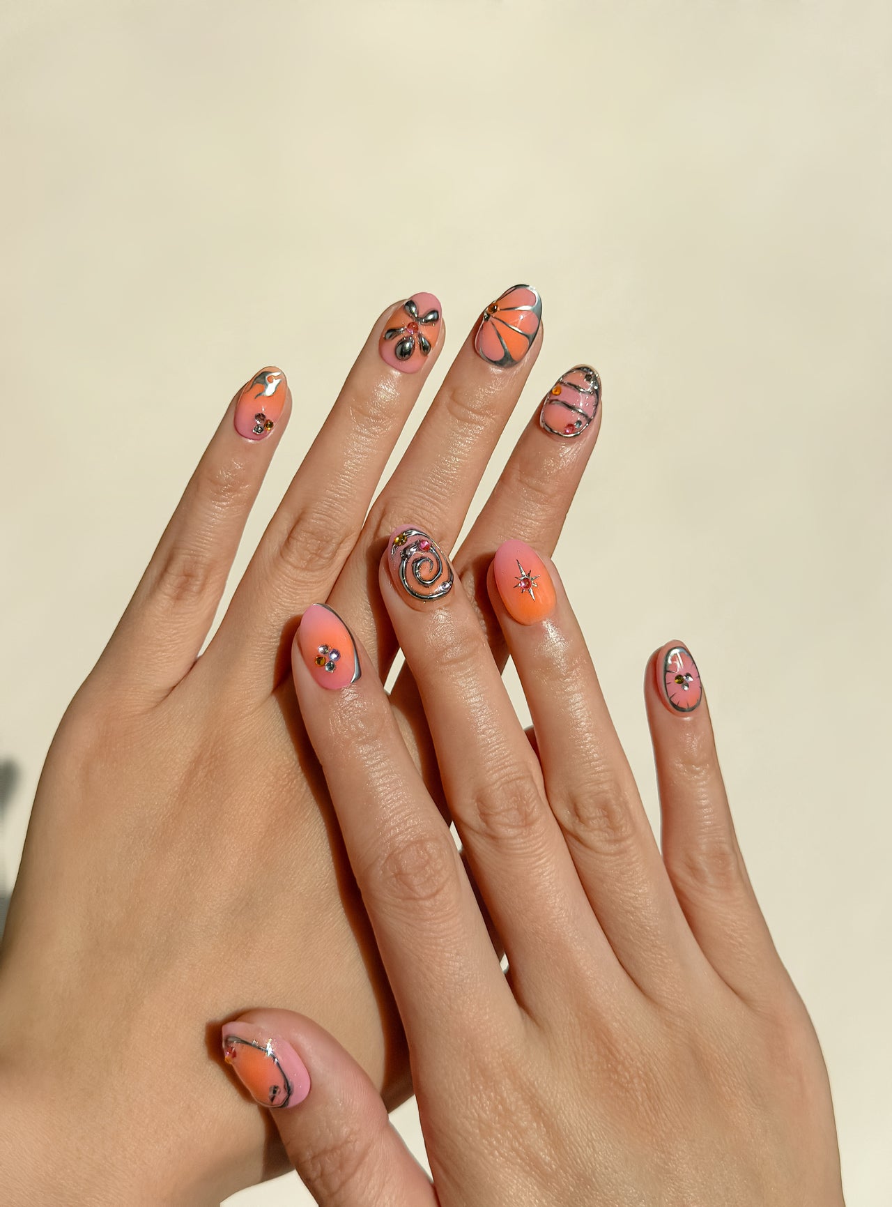 Coral Crush handmade reusable press-on nails laid flat, featuring coral and pink ombré nail art, chrome outlines, abstract swirls, floral motifs and gem accents in a extra short oval shape.