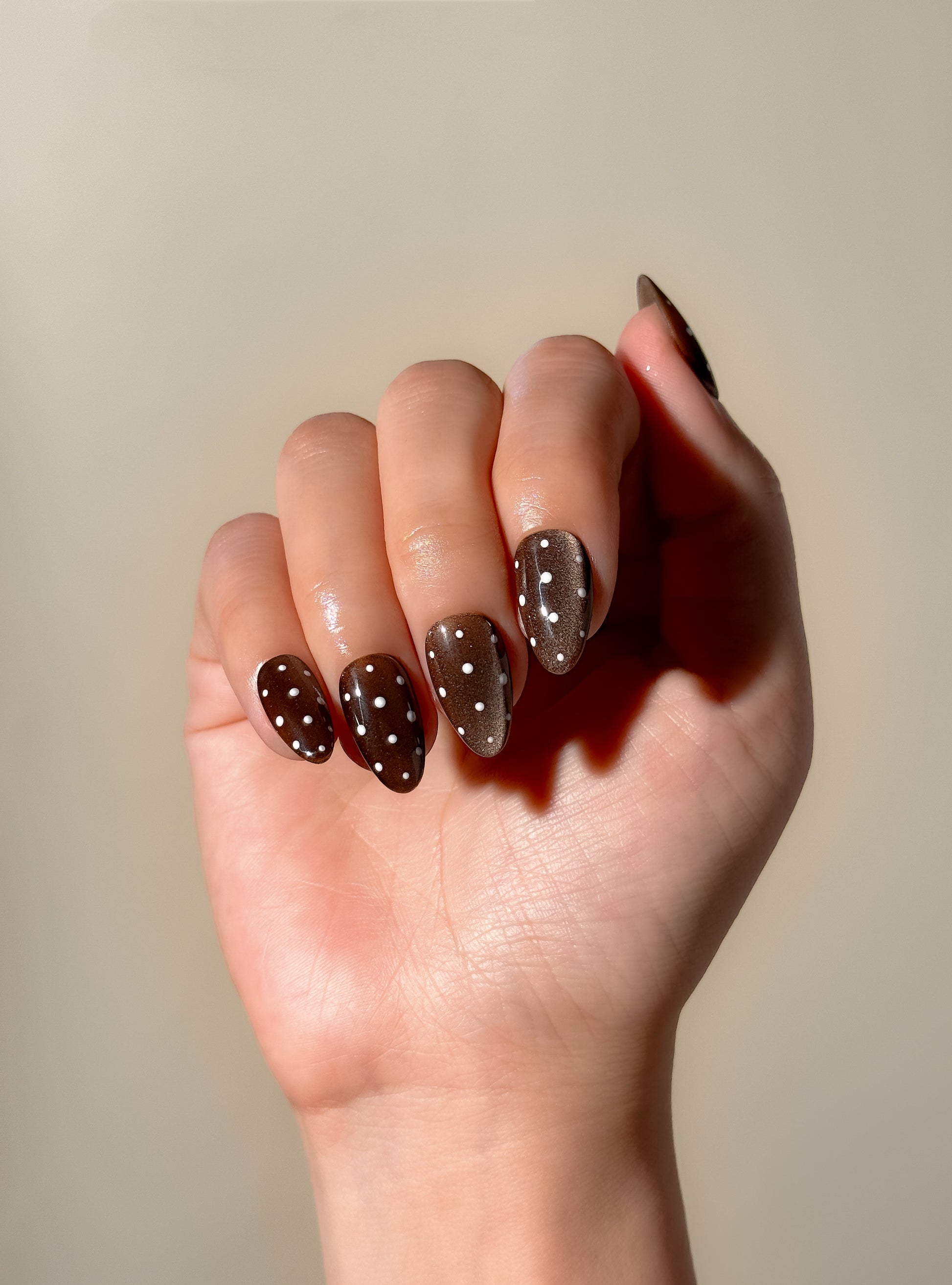Mocha Dot press-on nails worn on one hand, showing a rich brown cat-eye finish with white polka dots and a glossy medium almond shape.
