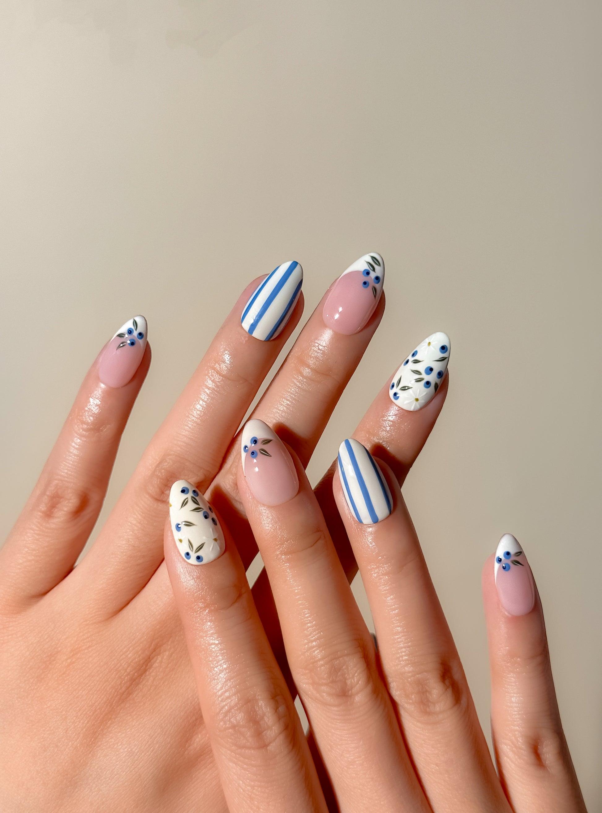 Blue and white Mediterranean-inspired press-on nails with hand-painted floral detailing and striped accents in a short almond shape. Handmade reusable false nails inspired by European summer nails, coastal Italian nail art and vintage ceramic patterns.