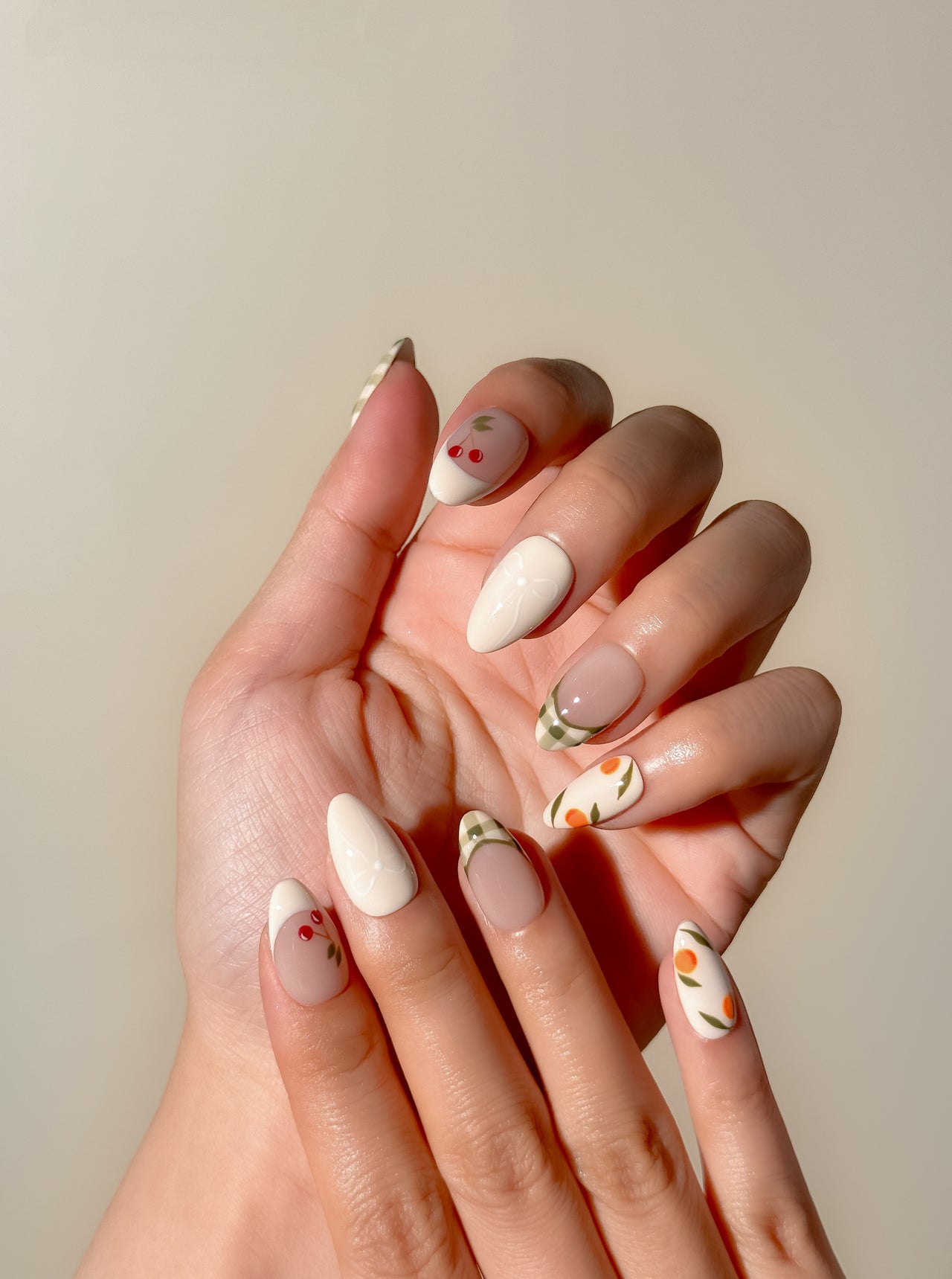 Cream and olive green press-on nails with hand-painted cherry, citrus and gingham French tip detailing in a medium almond shape. Handmade reusable false nails inspired by picnic aesthetics, European summer styling and vintage fruit nail art.