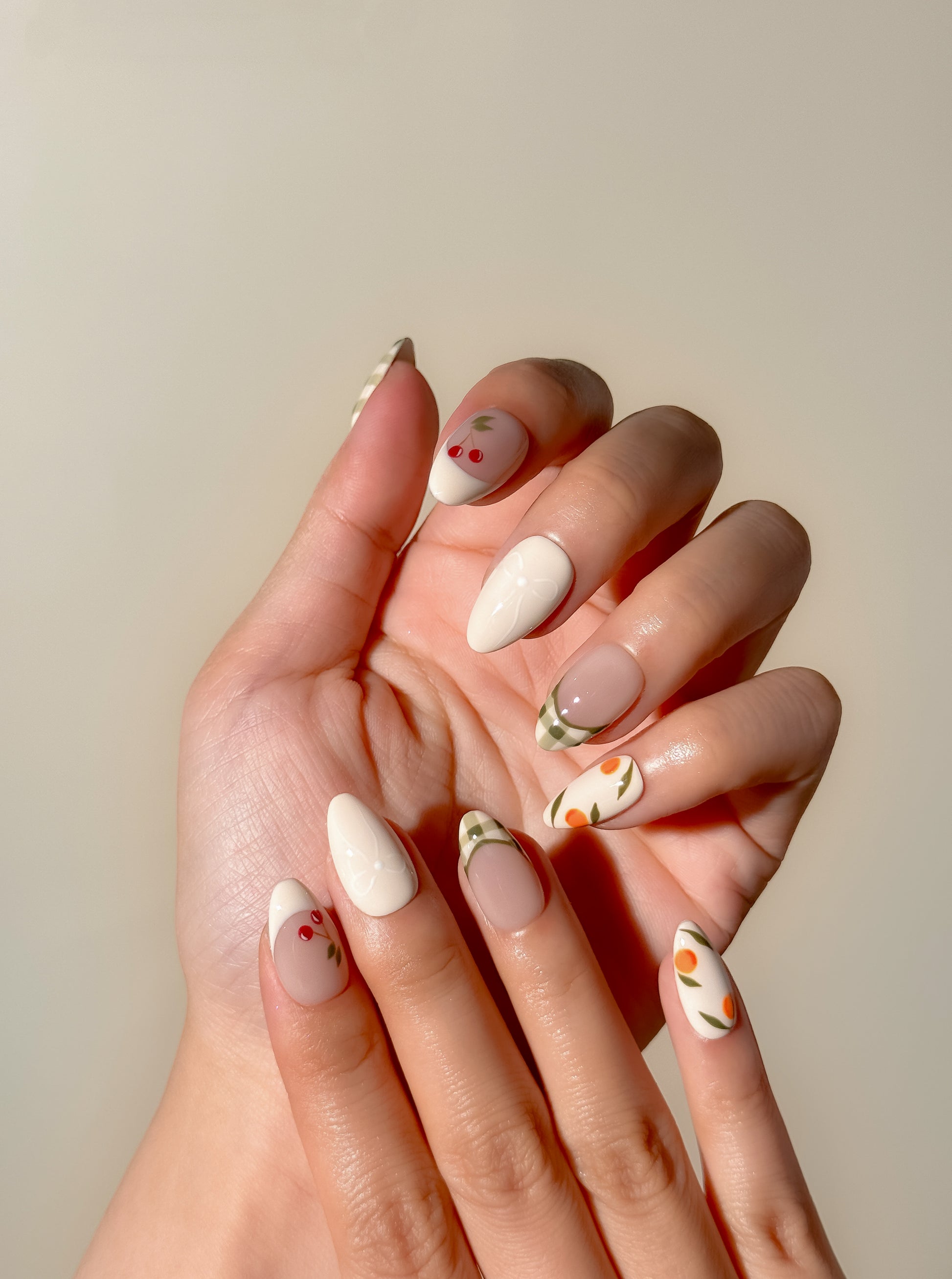 Cream and olive green press-on nails with hand-painted cherry, citrus and gingham French tip detailing in a medium almond shape. Handmade reusable false nails inspired by picnic aesthetics, European summer styling and vintage fruit nail art.