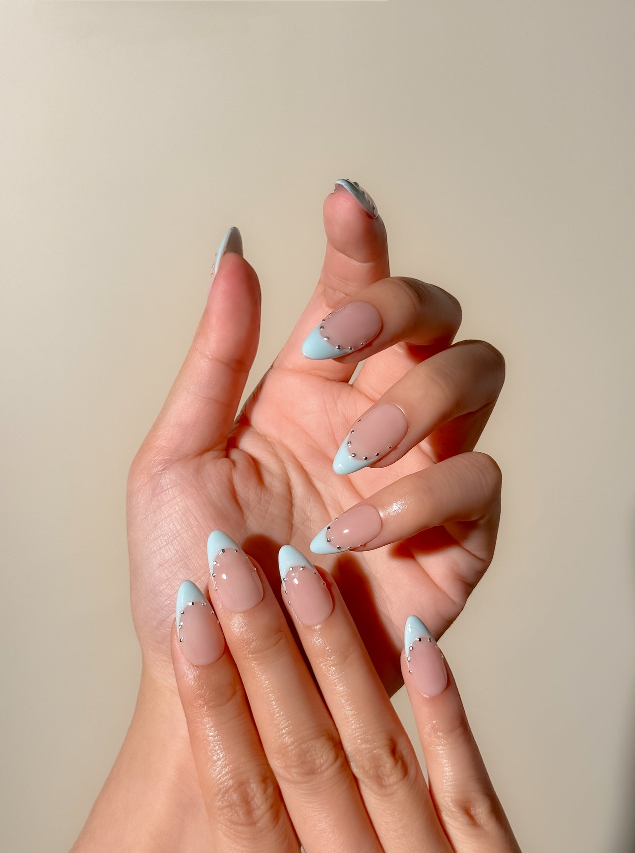 Powder blue French tip press-on nails with silver stud detailing in a glossy medium almond shape. Handmade reusable false nails with a sheer nude pink base inspired by European Riviera summer style and soft glam French manicure trends.