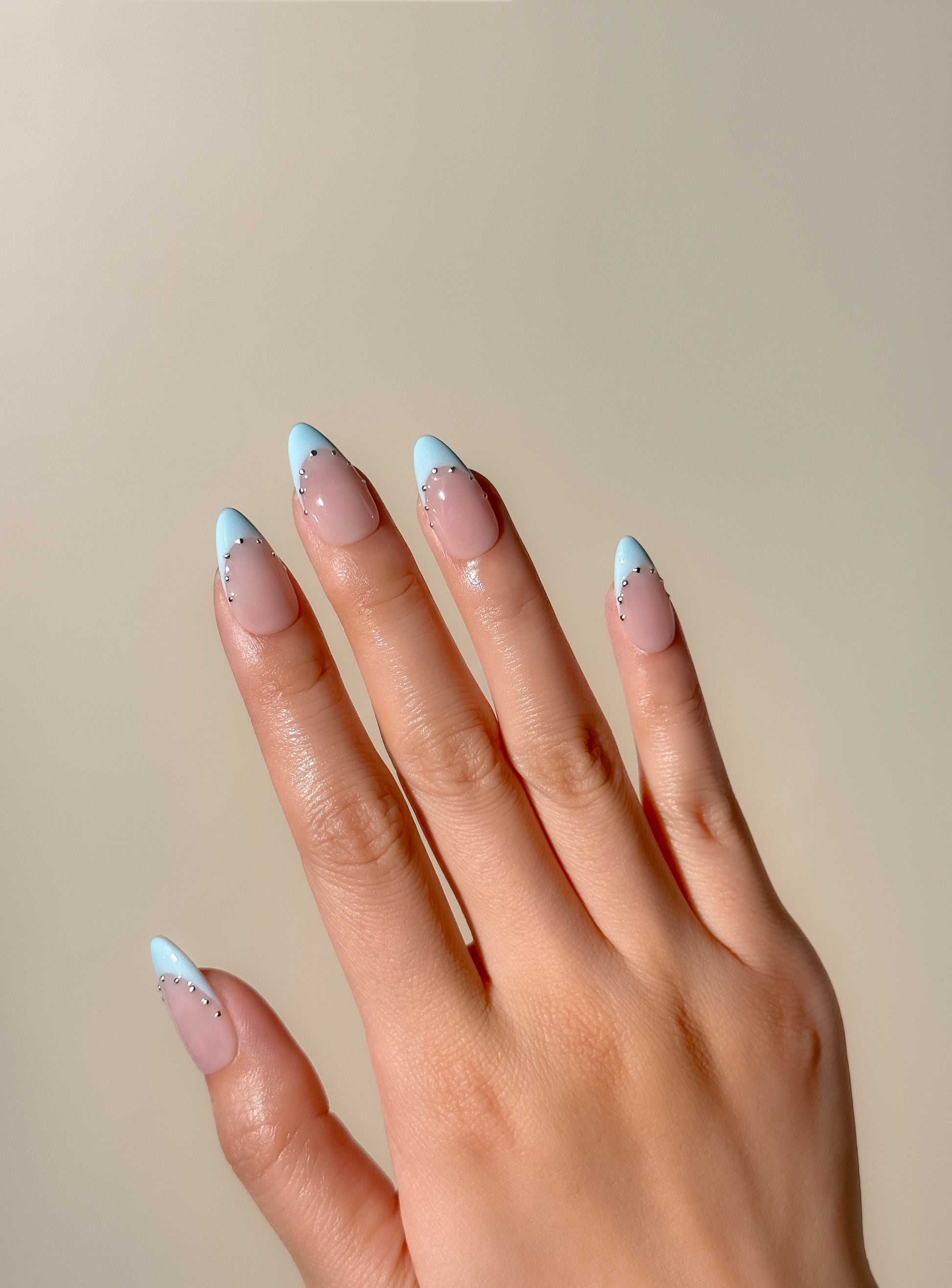 Soft blue French tip press-on nails with silver stud detailing in a glossy medium almond shape, photographed on hand in warm natural lighting. Nude pink base with icy powder blue tips inspired by European Riviera summer style.
