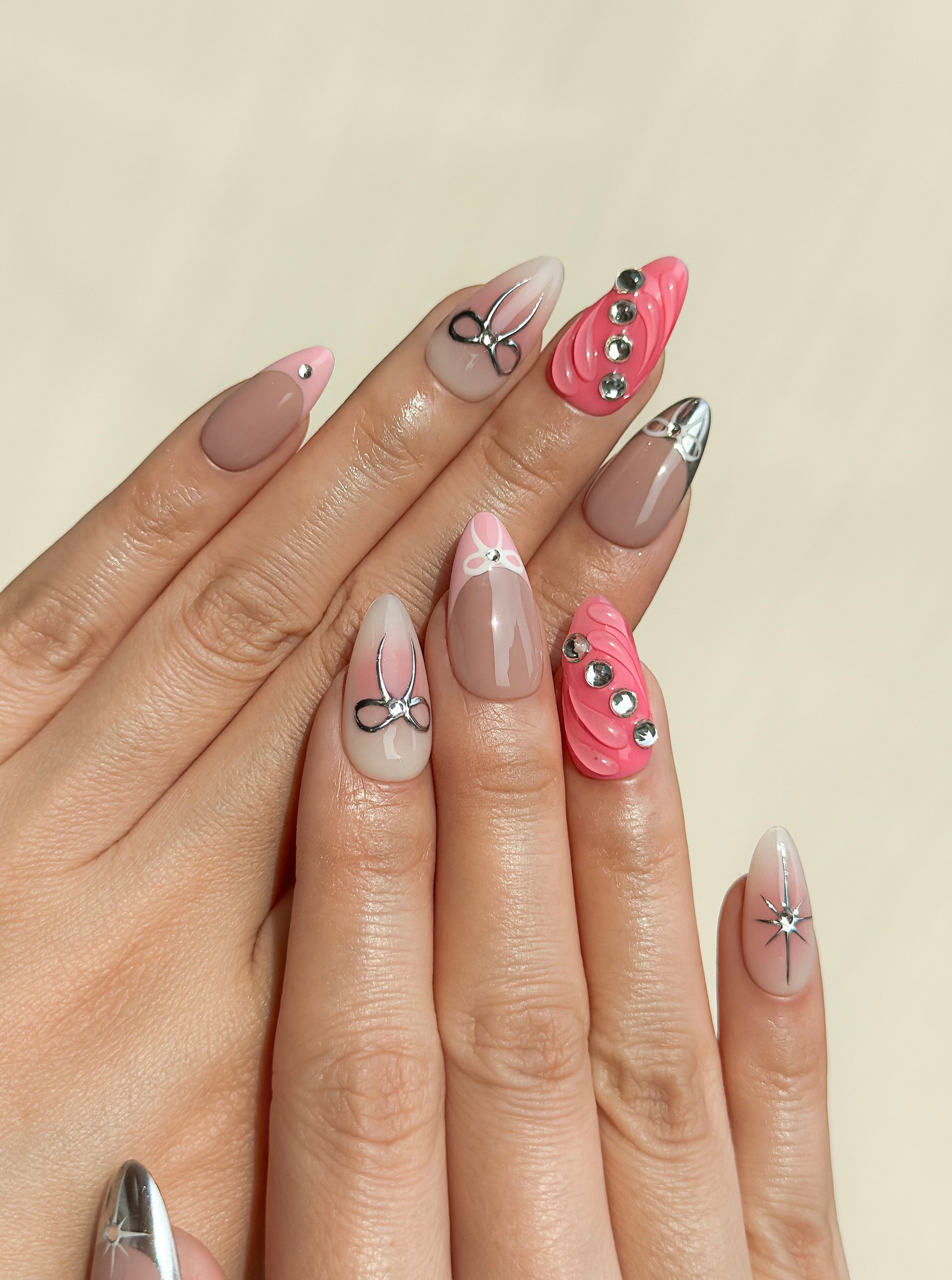 Starlit handmade press-on nails on hands, featuring silver chrome bows, crystal details, soft nude bases, star nail art and hot pink textured accent nails with a glossy finish.