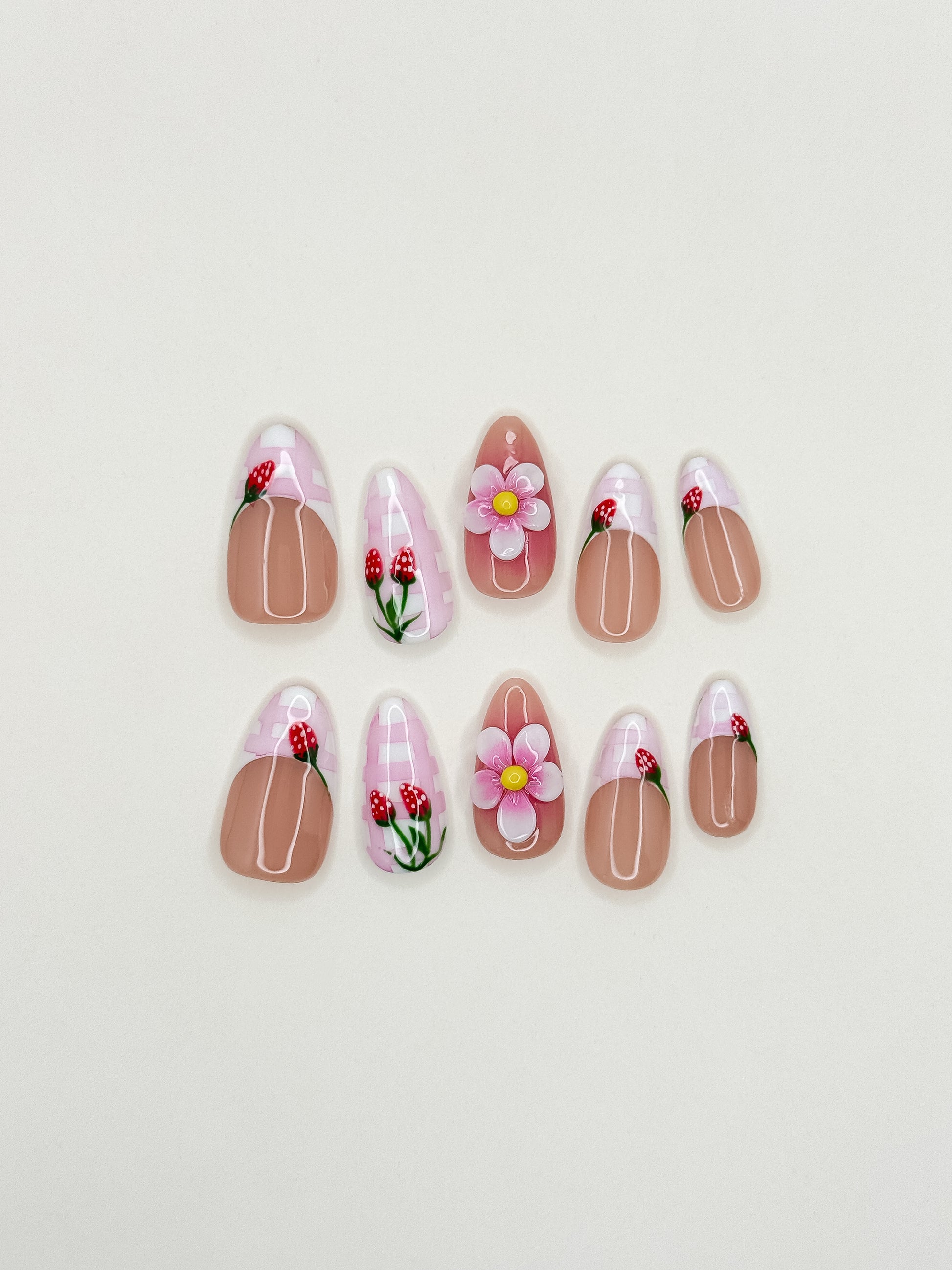 Strawberry Dream handmade reusable press-on nails laid flat, featuring sheer nude bases, pink gingham French tips, hand-painted strawberry details and raised 3D flower accents in a medium almond shape.