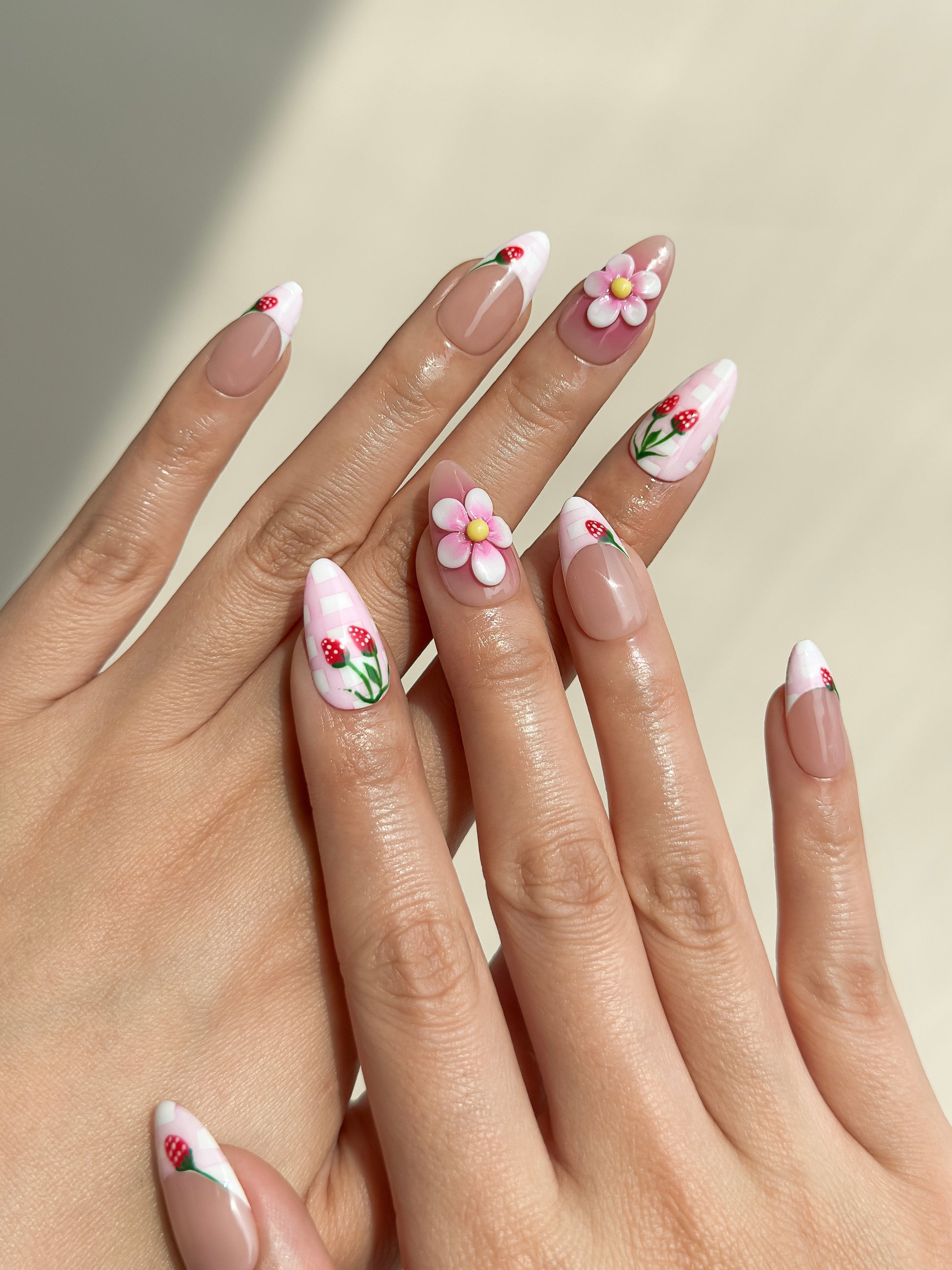 Strawberry Dream handmade press-on nails on hands, featuring pink plaid French tips, red strawberry details, sculpted pink and white flowers and glossy medium almond nails.