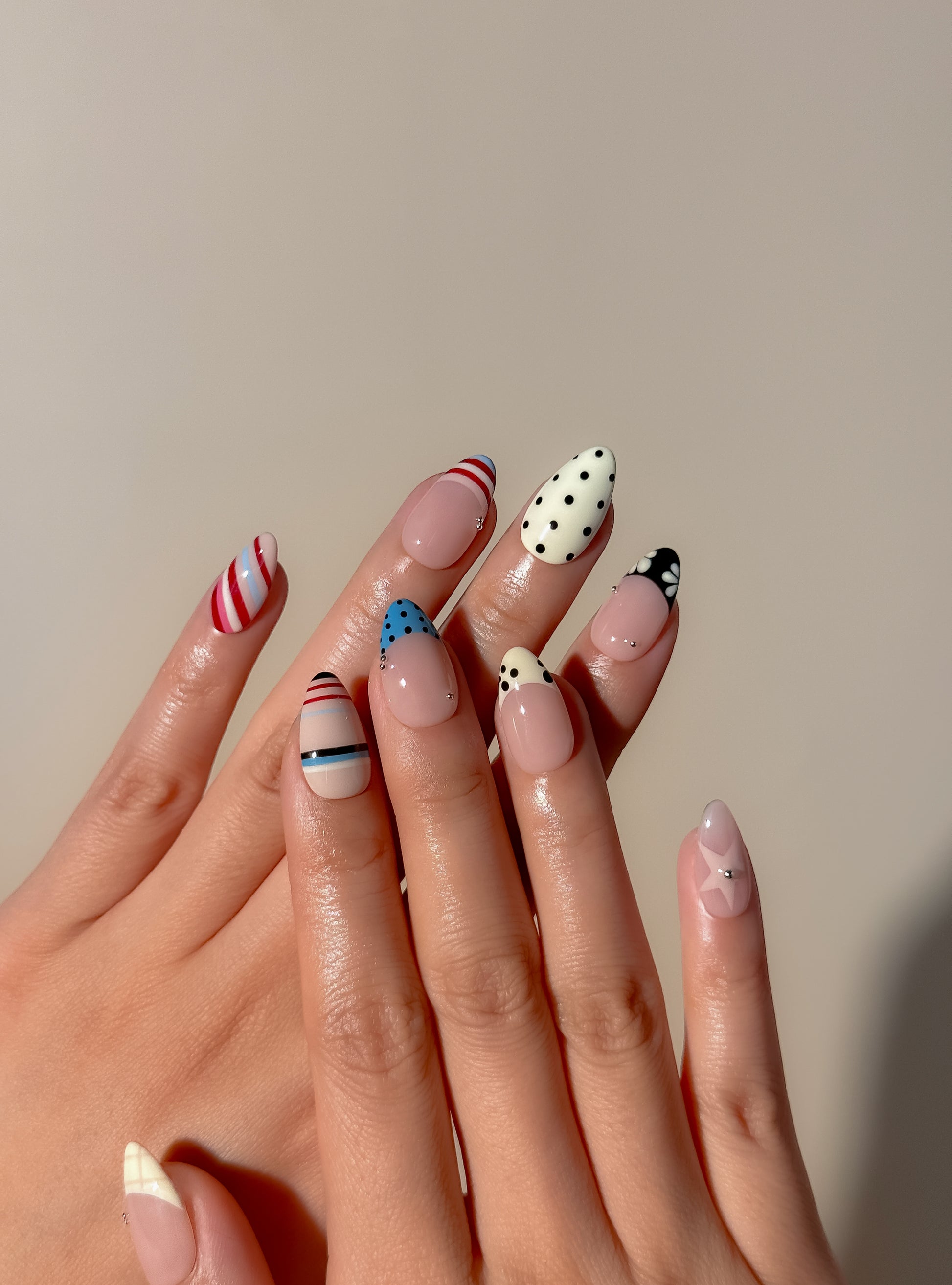 Colourful retro Stripe Club press-on nails worn on hands, with red and blue stripes, black polka dots, cream accents and glossy nude bases in a short almond shape.