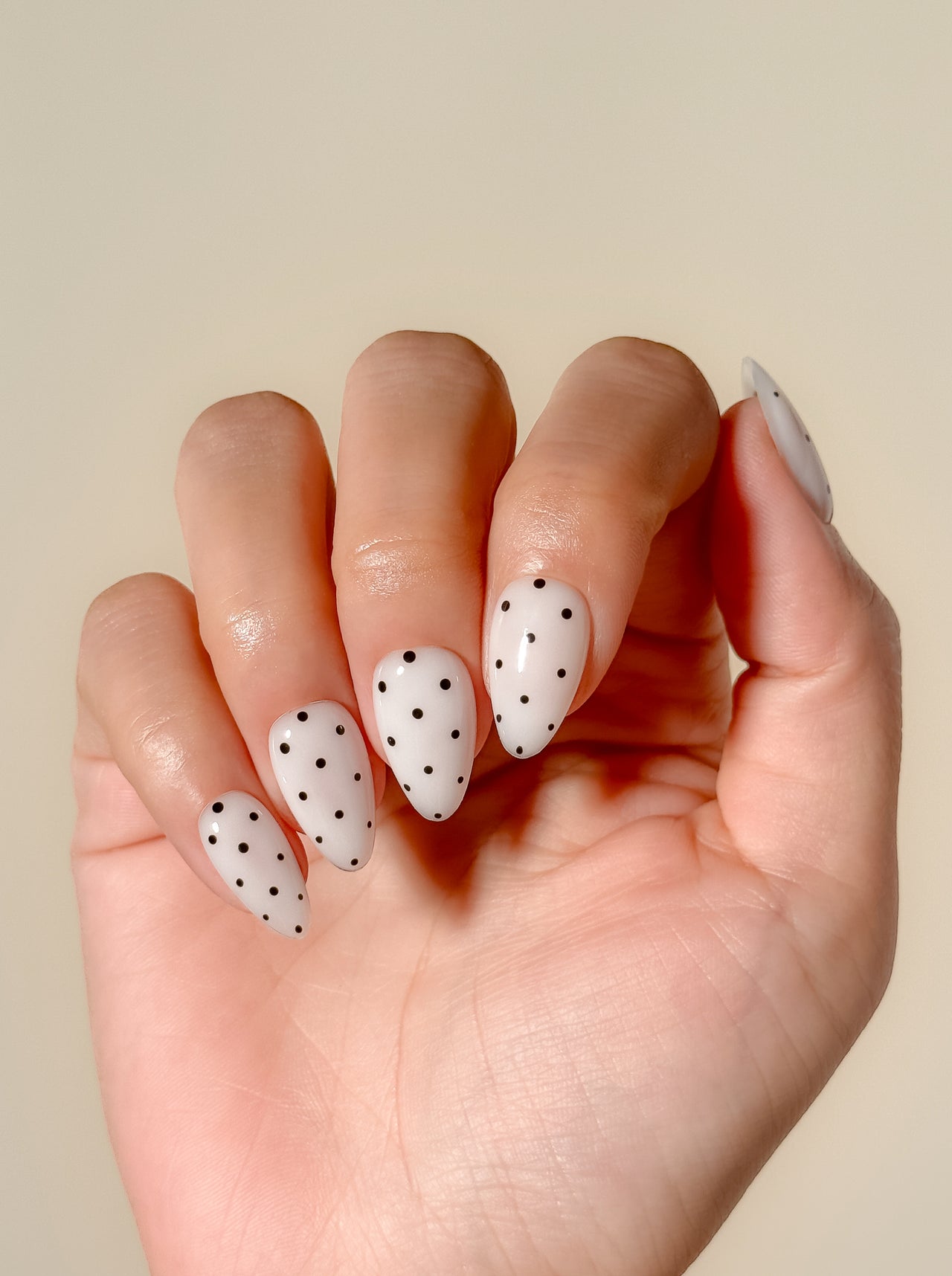 Cream white press-on nails with black polka dot nail art in a glossy short almond shape. Handmade reusable false nails inspired by retro mod fashion, monochrome minimalist nails and vintage polka dot manicure styles.