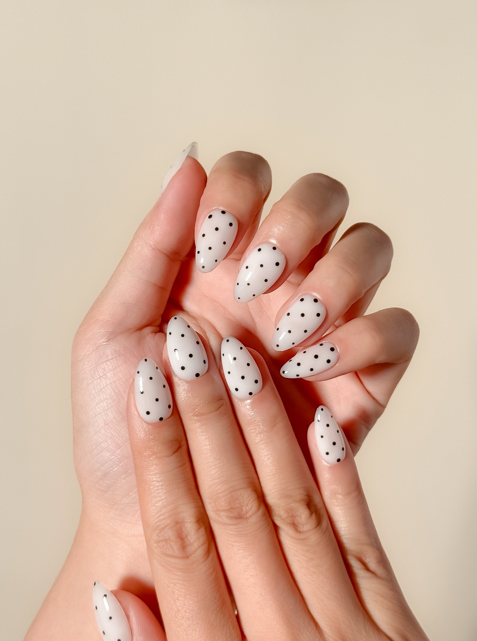 Cream white press-on nails with black polka dot nail art in a glossy short almond shape. Handmade reusable false nails inspired by retro mod fashion, monochrome minimalist nails and vintage polka dot manicure styles.