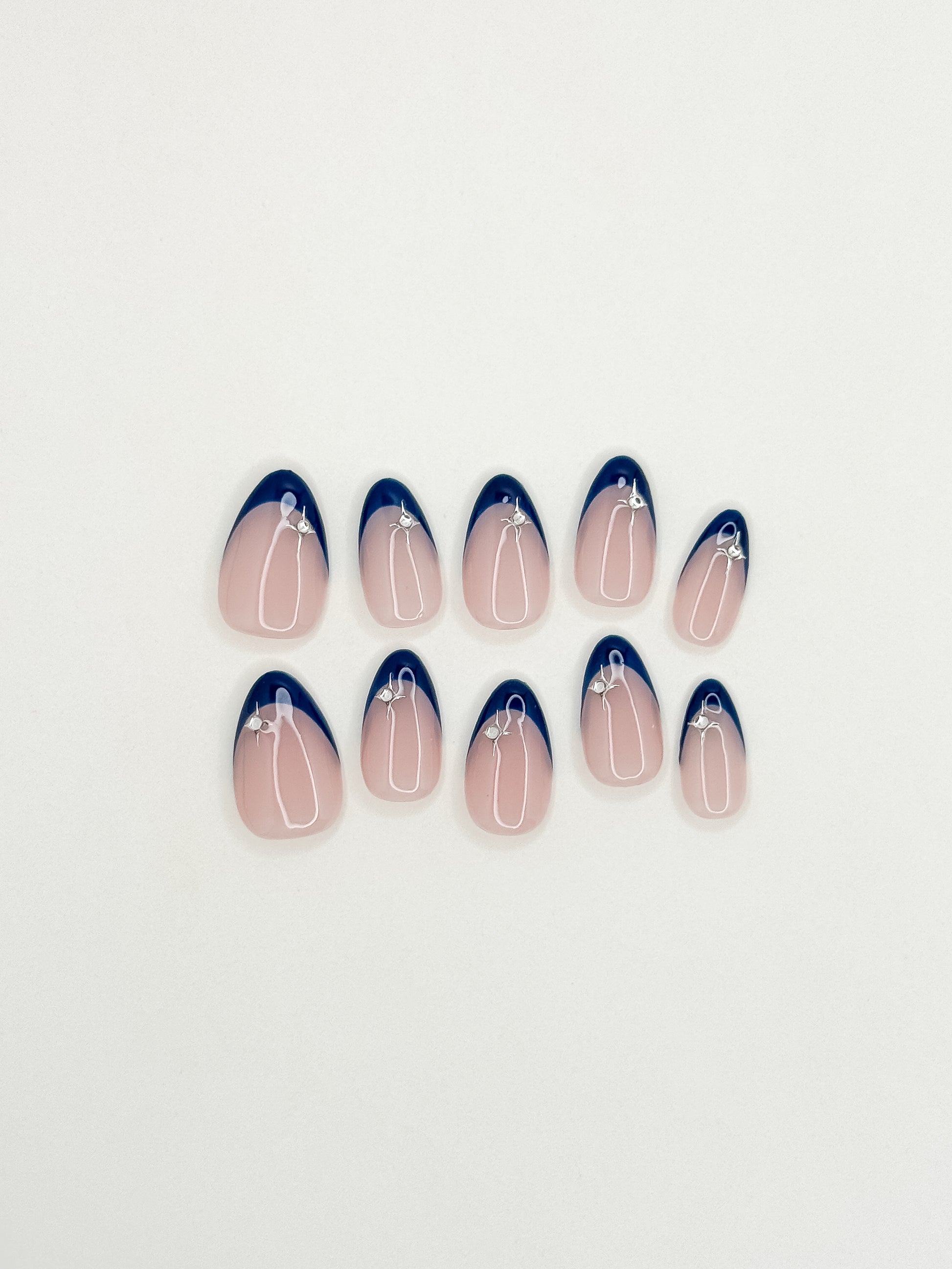 After Midnight handmade press-on nails arranged on a white background, featuring short almond nails with deep navy skinny French tips, a sheer blush base and silver star embellishments.