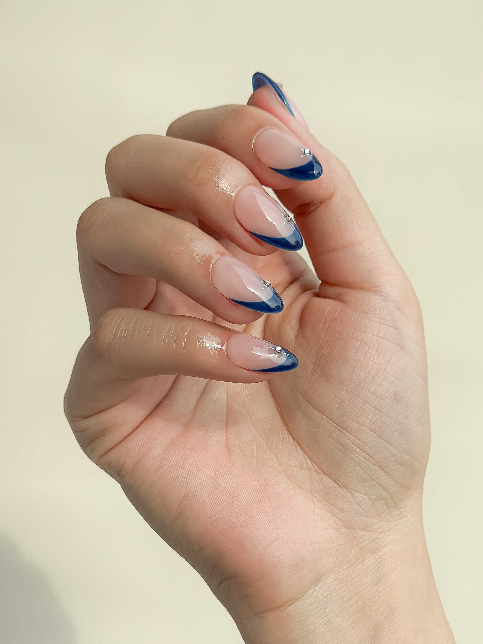 Close-up of After Midnight short almond press-on nails worn on hand, with glossy navy French tips, a sheer blush-pink base and silver celestial embellishments.