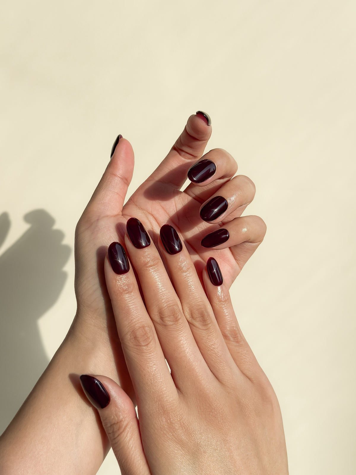 Two hands wearing After Work glossy dark cherry burgundy short oval press-on nails in warm natural sunlight.