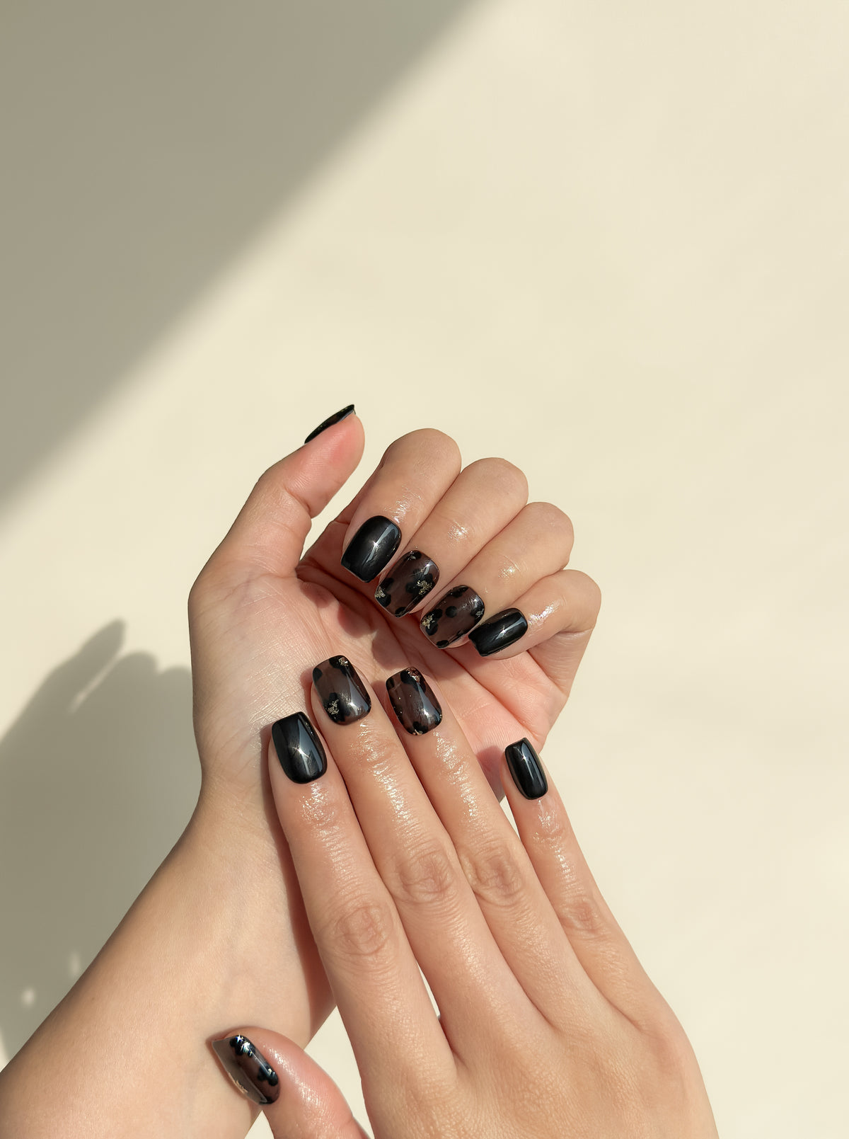 Two hands wearing Allegedly smoky black abstract short square press-on nails with high-gloss gold foil details.