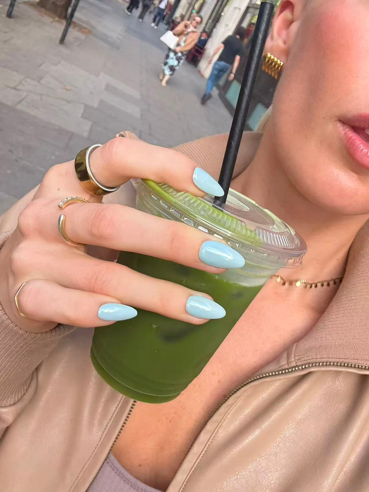 A close-up lifestyle shot of a person holding a green iced drink in a clear plastic cup with a black straw. The person has long, light blue almond-shaped nails, gold rings, and is wearing a beige leather jacket.