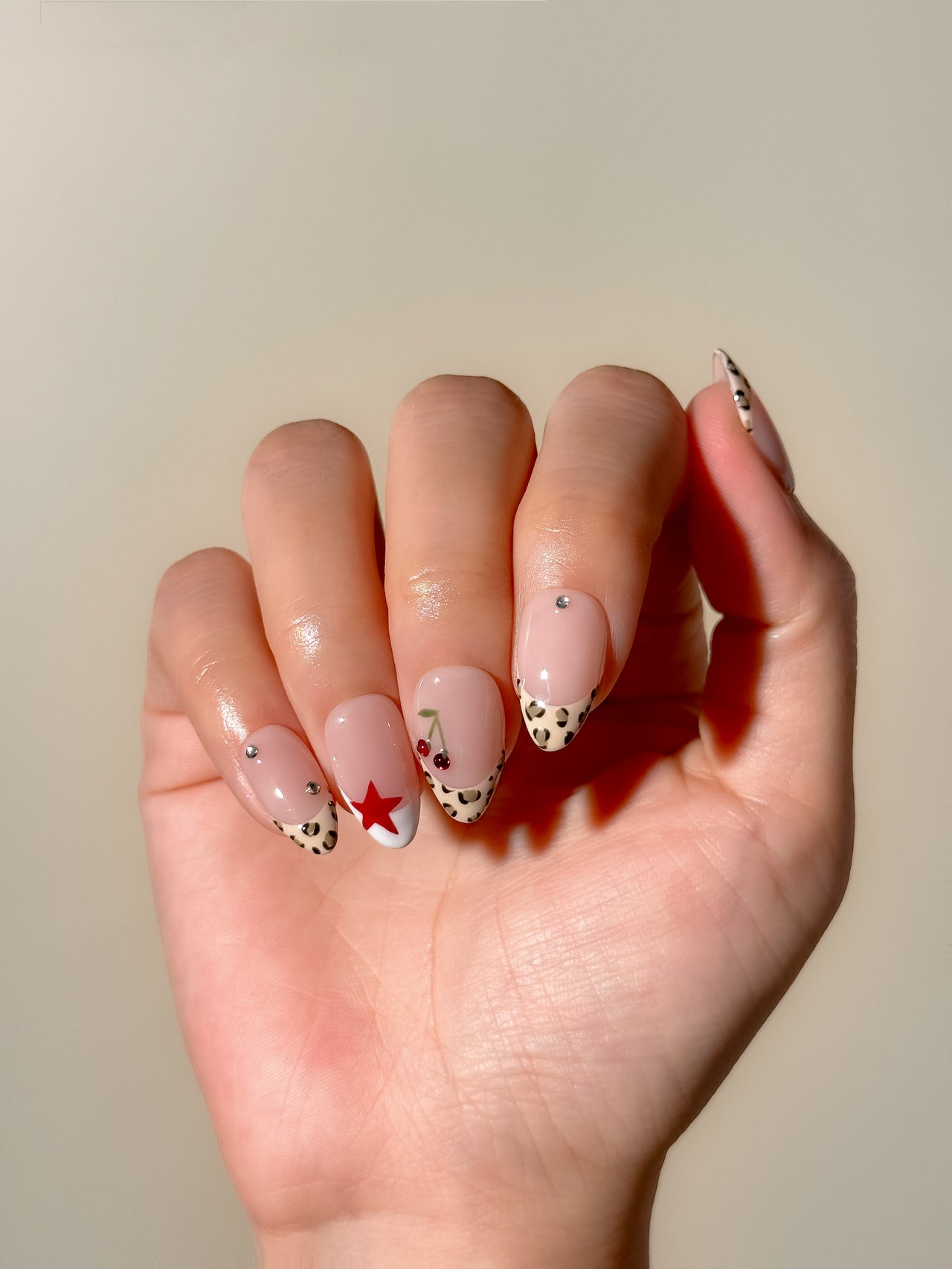 Cherry Bombshell handmade press-on nails arranged on a white background, featuring short almond nails with leopard-print skinny French tips, red cherries, red stars and crystal accents.