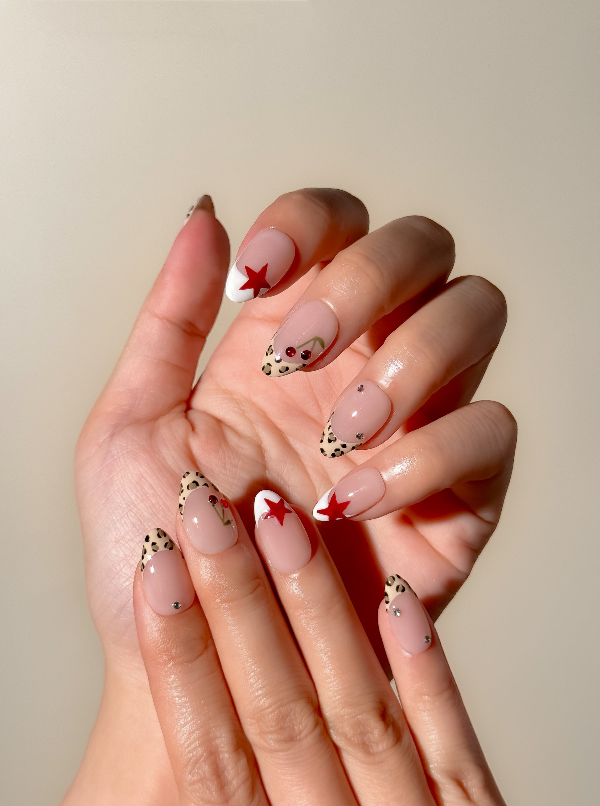 Cherry Bombshell handmade press-on nails worn on two hands, featuring short almond leopard French tips, red cherry and star accents, white detailing and crystals.