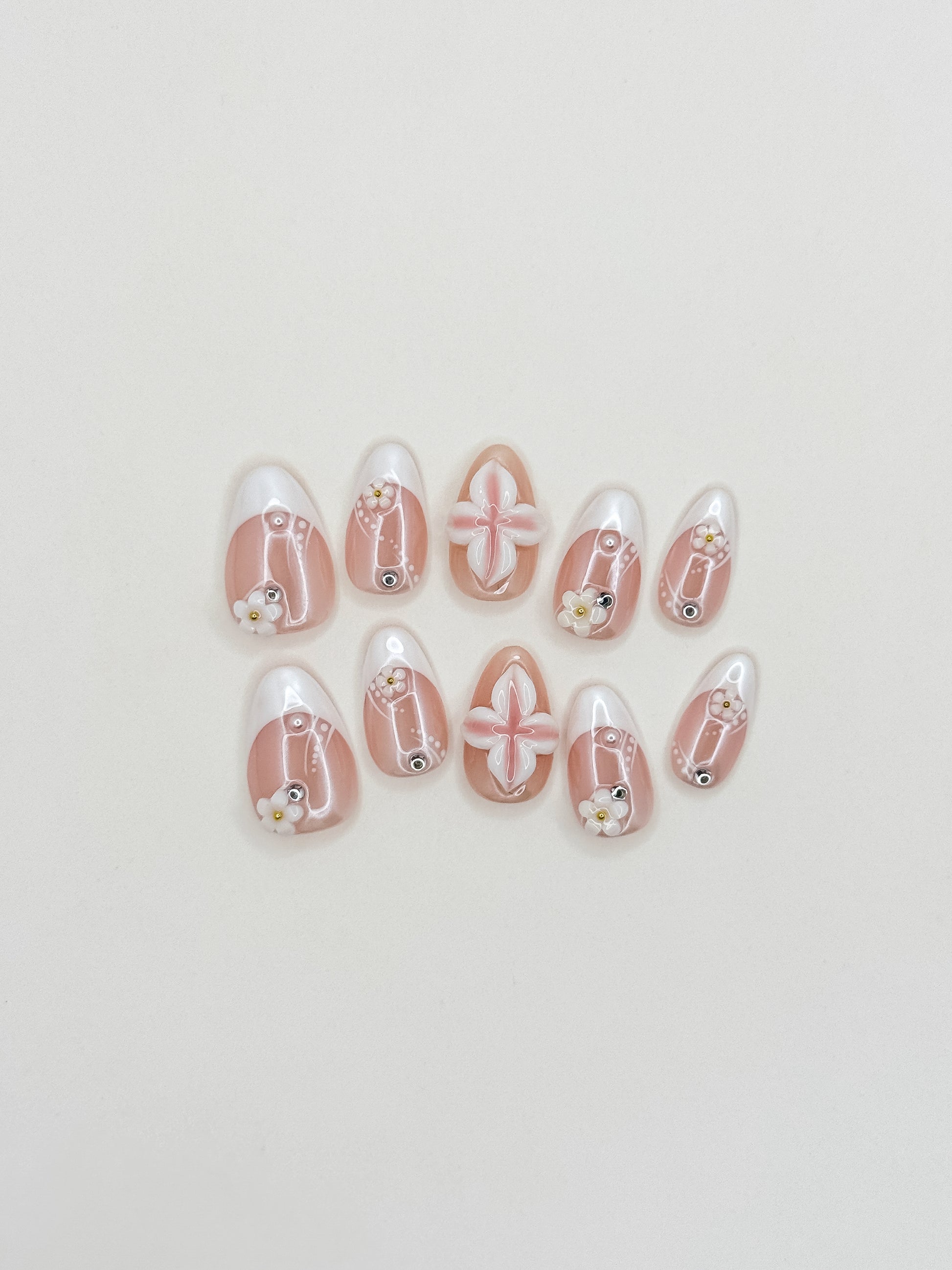 Last Call short almond press-on nails arranged on white, with sheer blush bases, glossy white French tips, 3D flowers and silver studs.