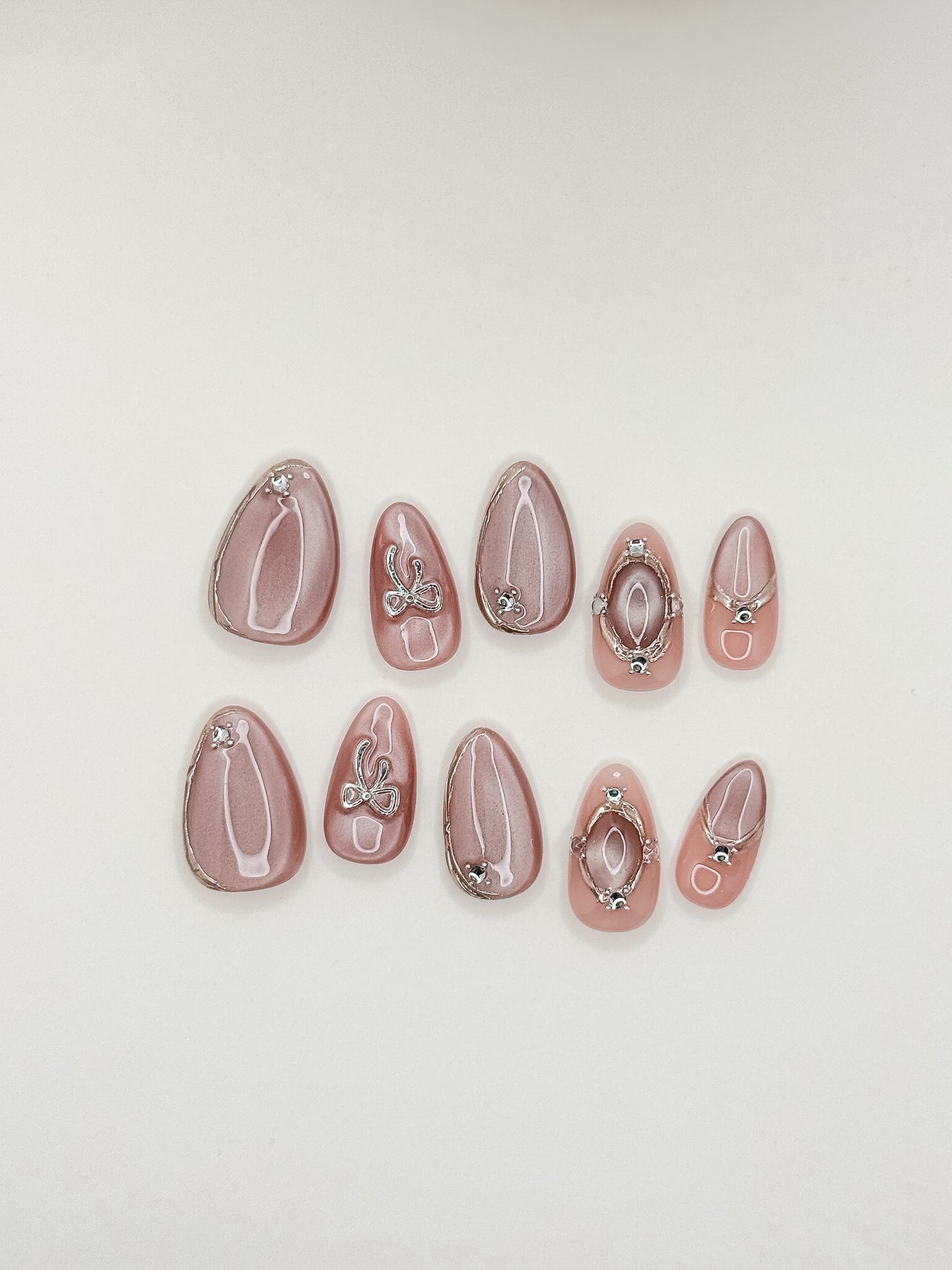 Late Checkout short almond press-on nails arranged on white, with rose-pink cat-eye shimmer, sculptural silver loops and crystal details.