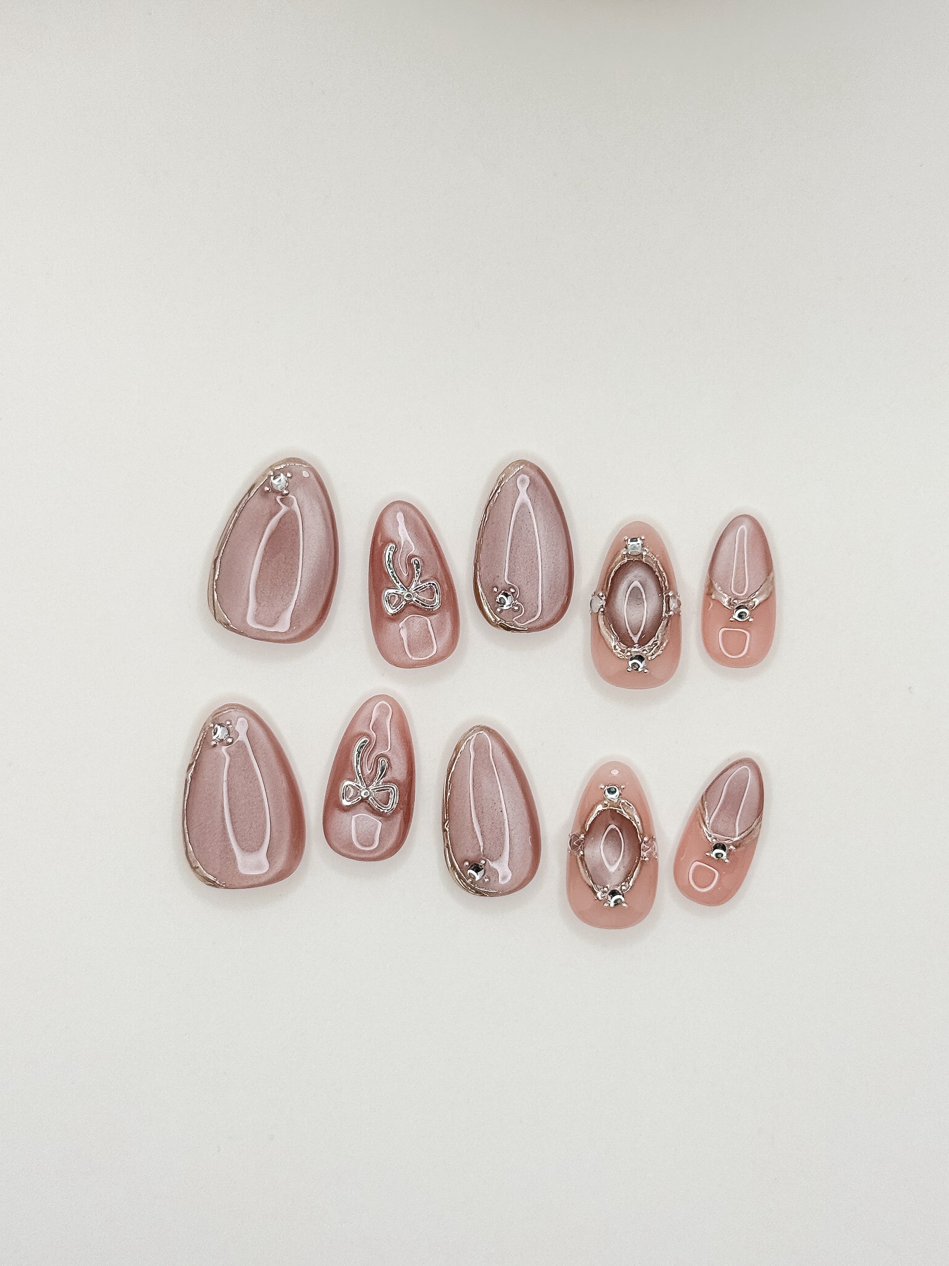 Late Checkout short almond press-on nails arranged on white, with rose-pink cat-eye shimmer, sculptural silver loops and crystal details.