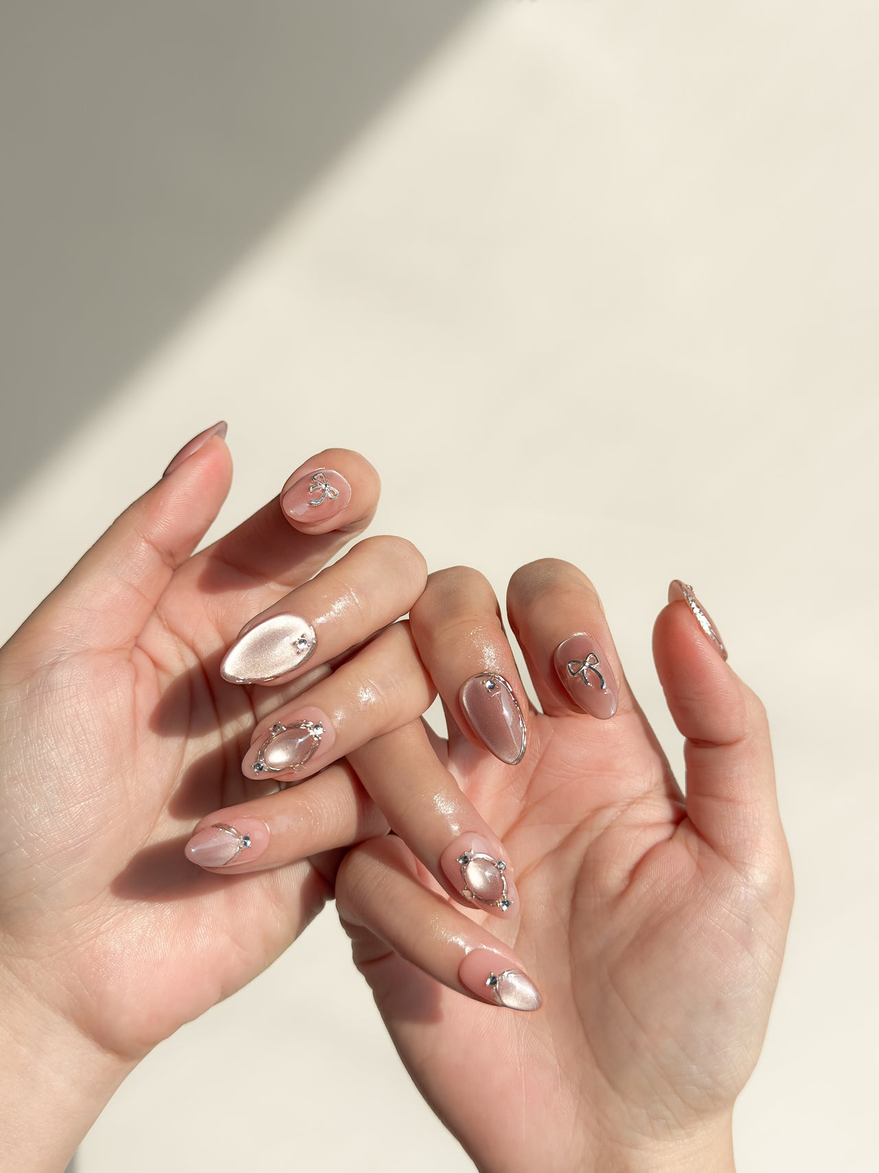 Late Checkout short almond press-on nails arranged on white, with rose-pink cat-eye shimmer, sculptural silver loops and crystal details.
