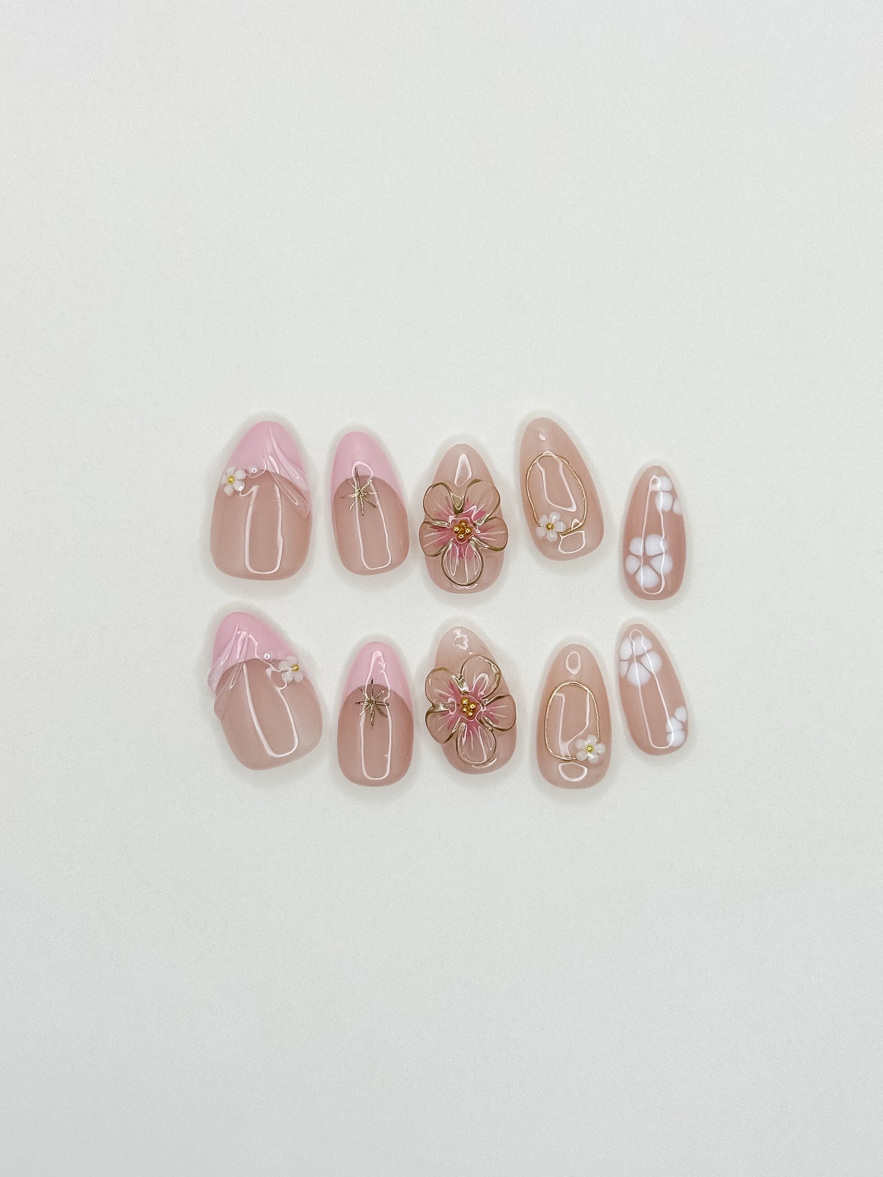 Petal handmade reusable press-on nails laid flat, featuring blush-pink bases, raised 3D flowers, white petals and gold details in a short almond shape.