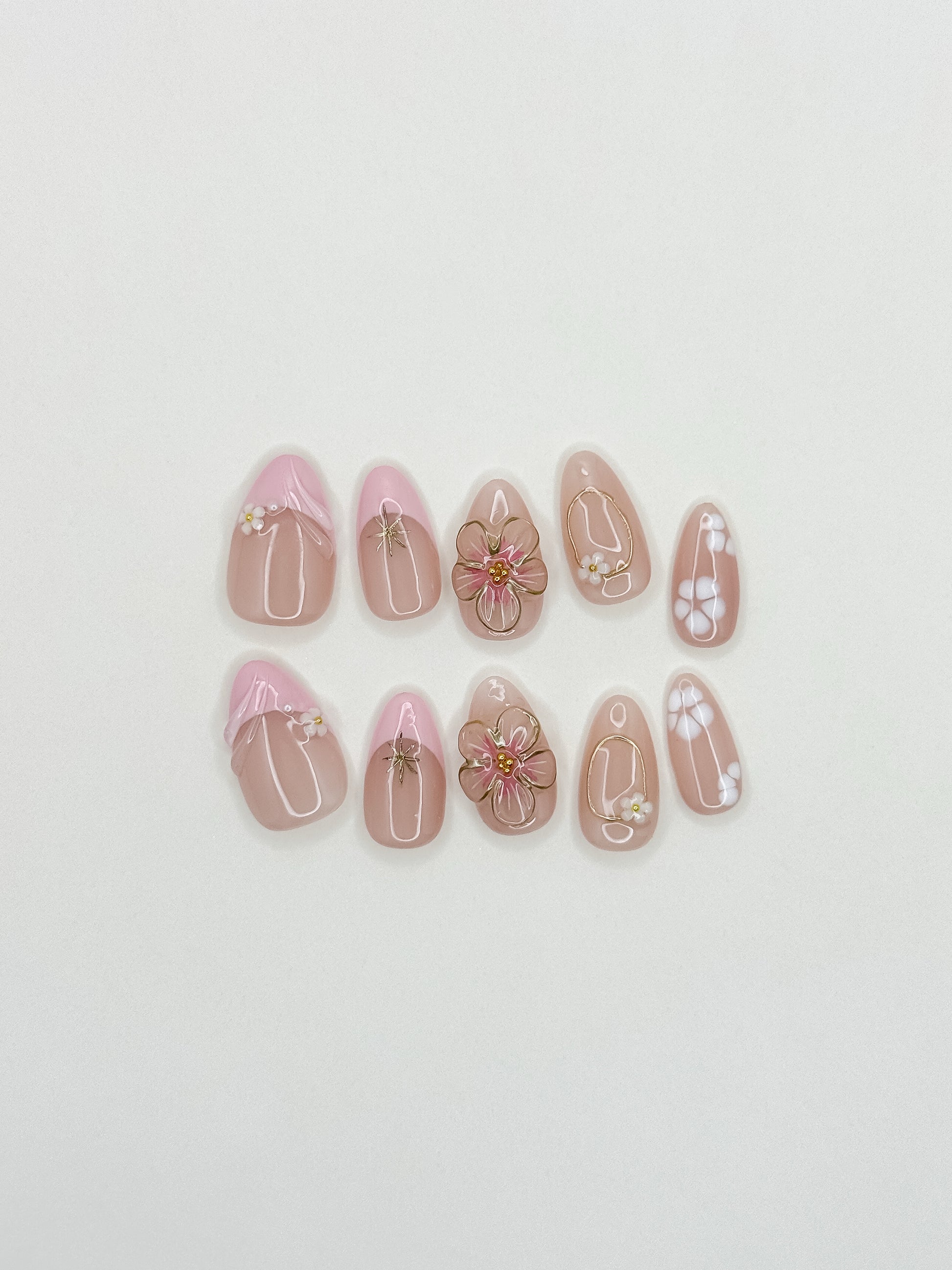 Petal handmade reusable press-on nails laid flat, featuring blush-pink bases, raised 3D flowers, white petals and gold details in a short almond shape.