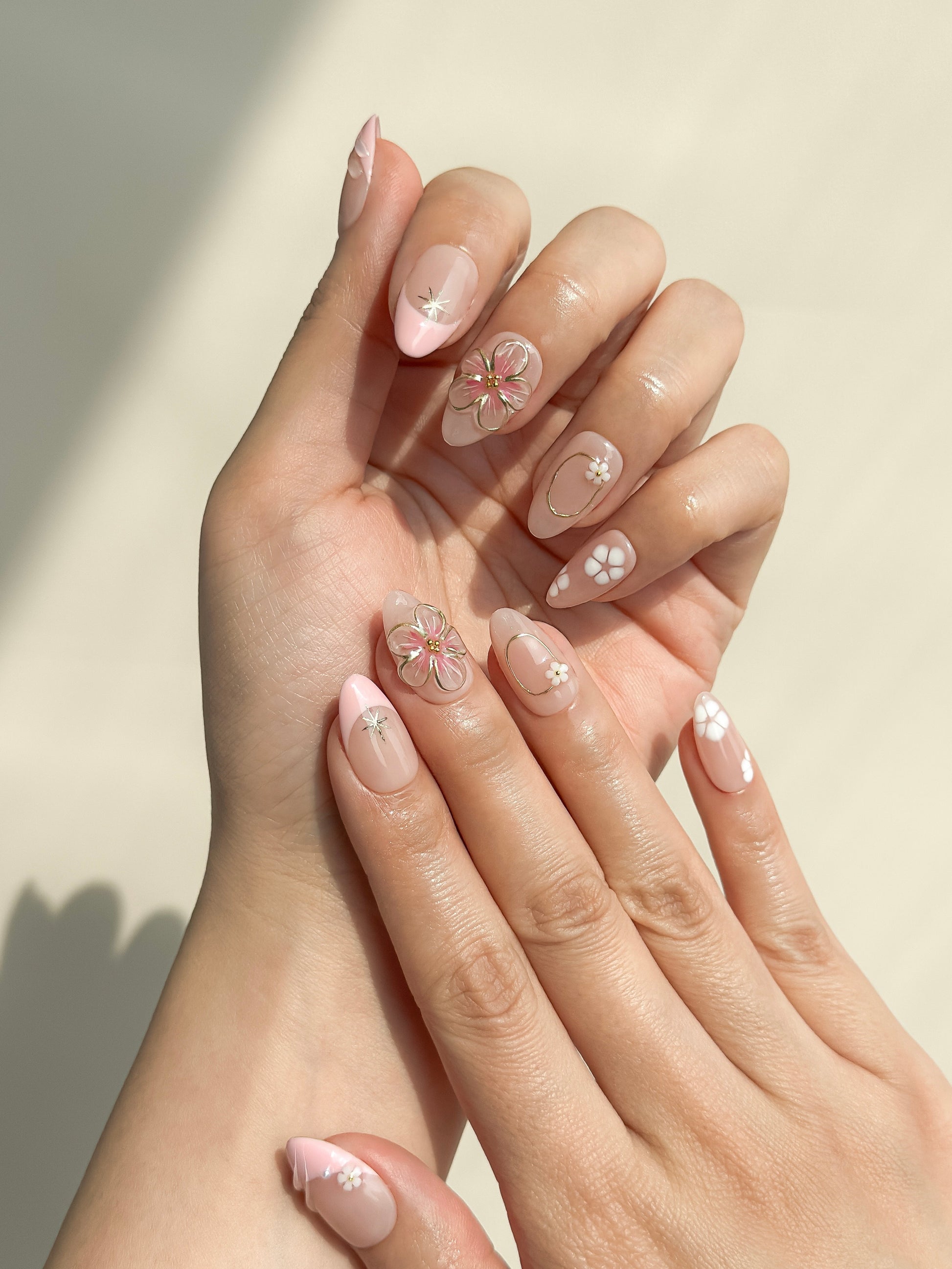 Petal pink press-on nails worn on both hands, showing sculpted 3D flowers, white floral accents, gold details and a glossy short almond shape.