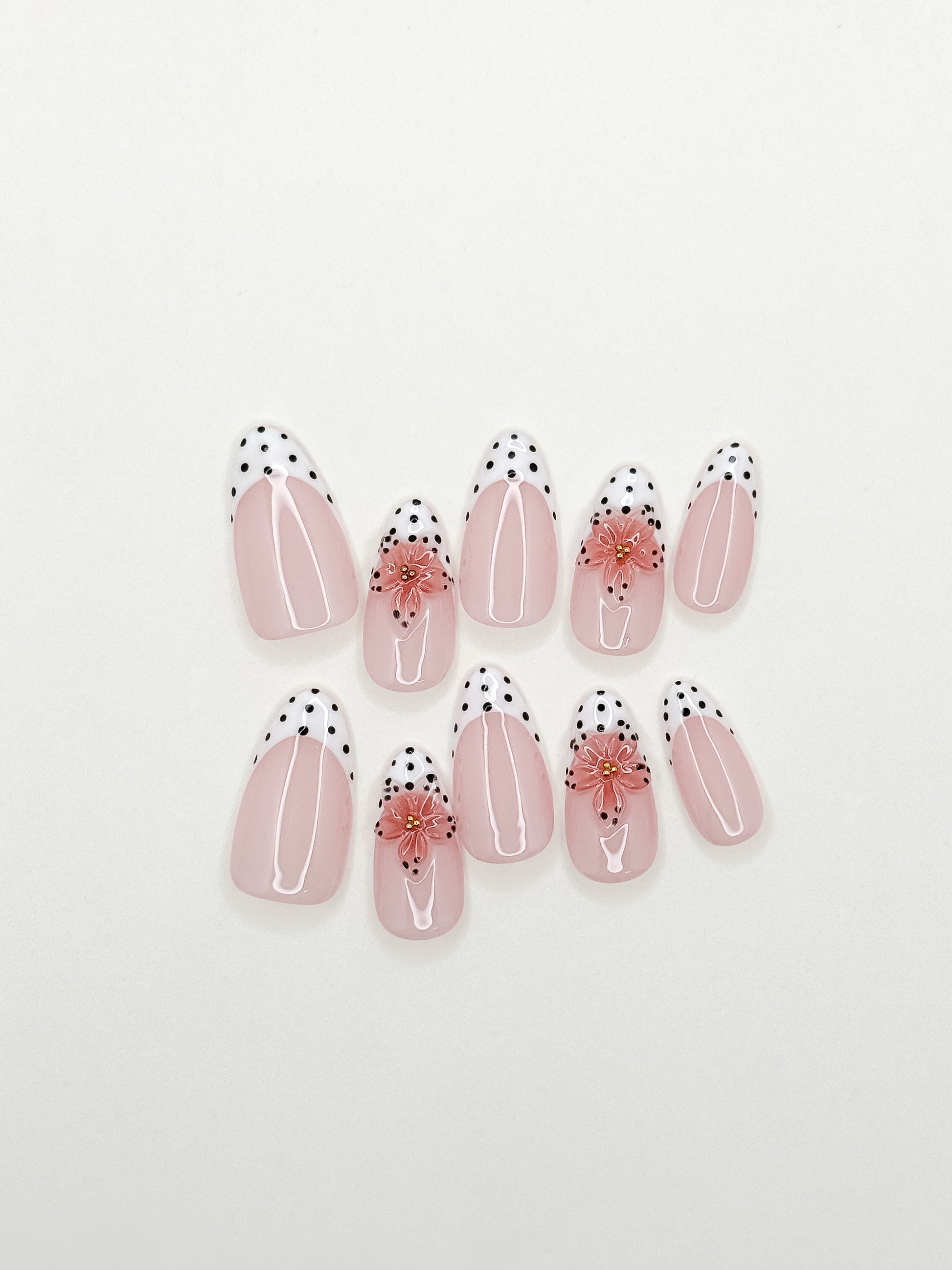 A set of ten false press-on nails arranged on a plain white background. The nails feature a glossy pale pink base with white curved stripes, black polka dots along the tips, and a small pink floral accent on two of the nails.