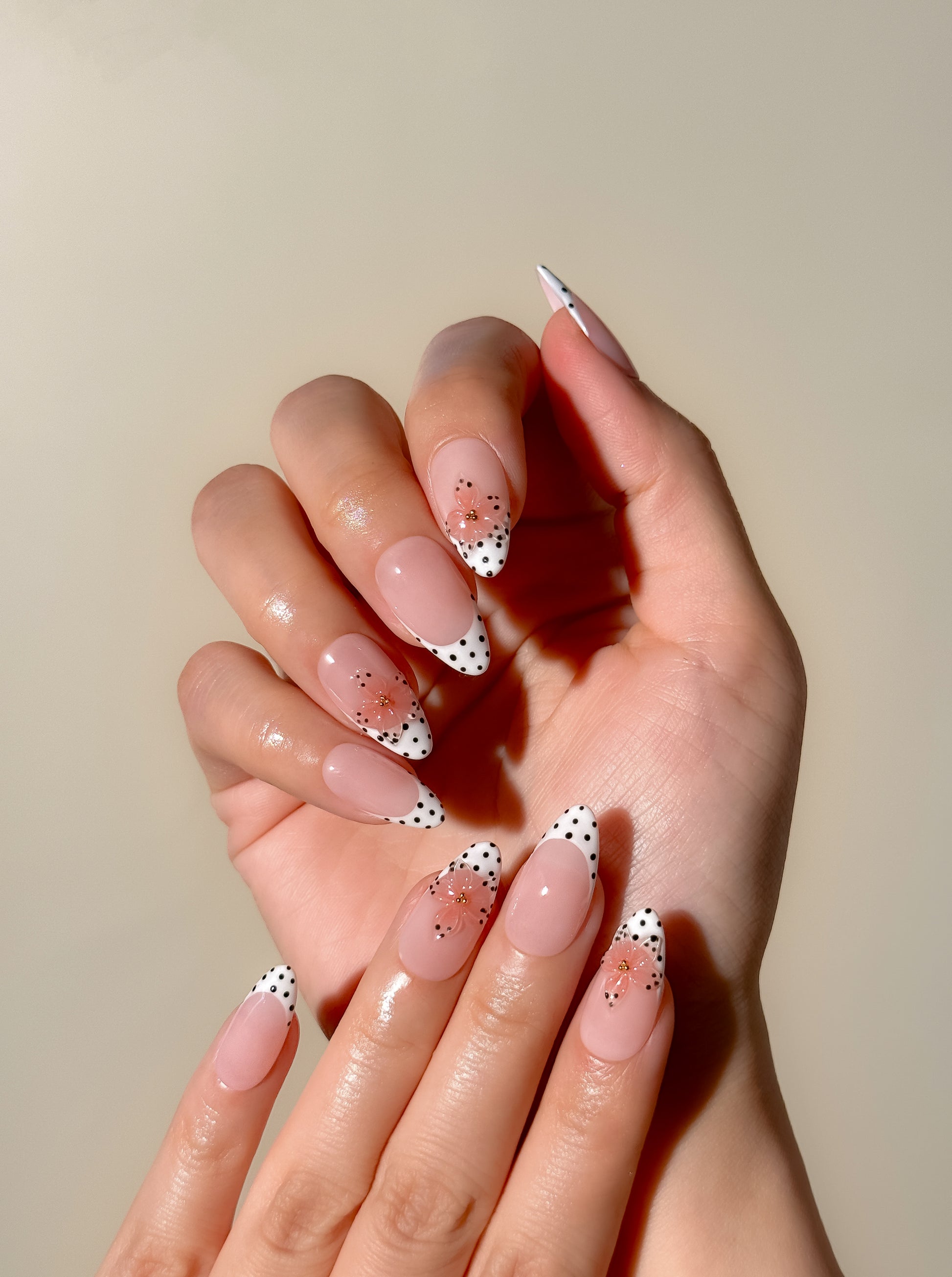 Polka Bloom medium almond press-on nails worn on two hands, featuring a sheer blush-pink base, white French tips with black polka dots, and translucent pink 3D flowers with gold centres.