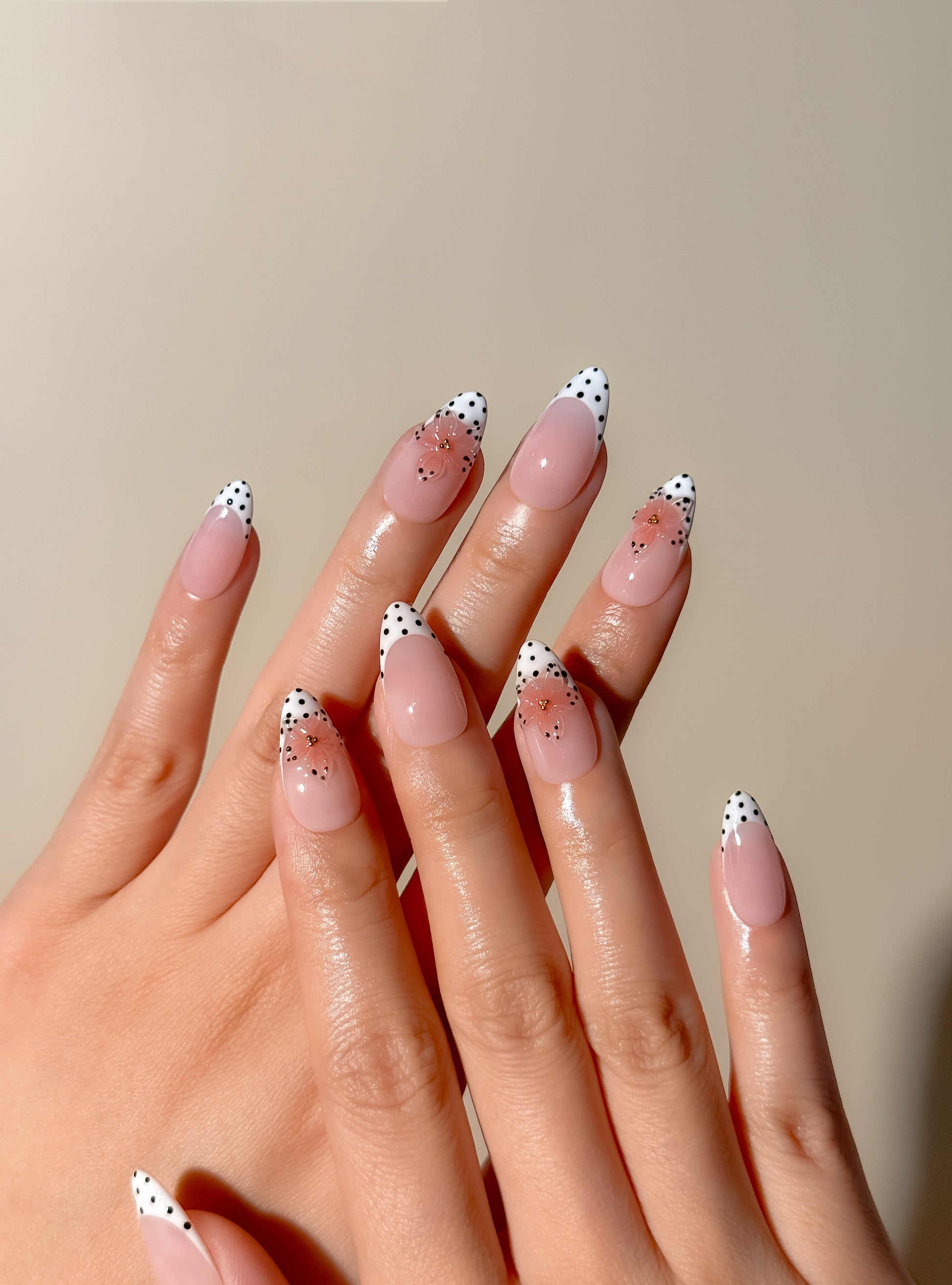 Polka Bloom medium almond press-on nails worn on two hands, with glossy nude-pink bases, black polka-dot French tips, and sculpted translucent pink floral nail art.