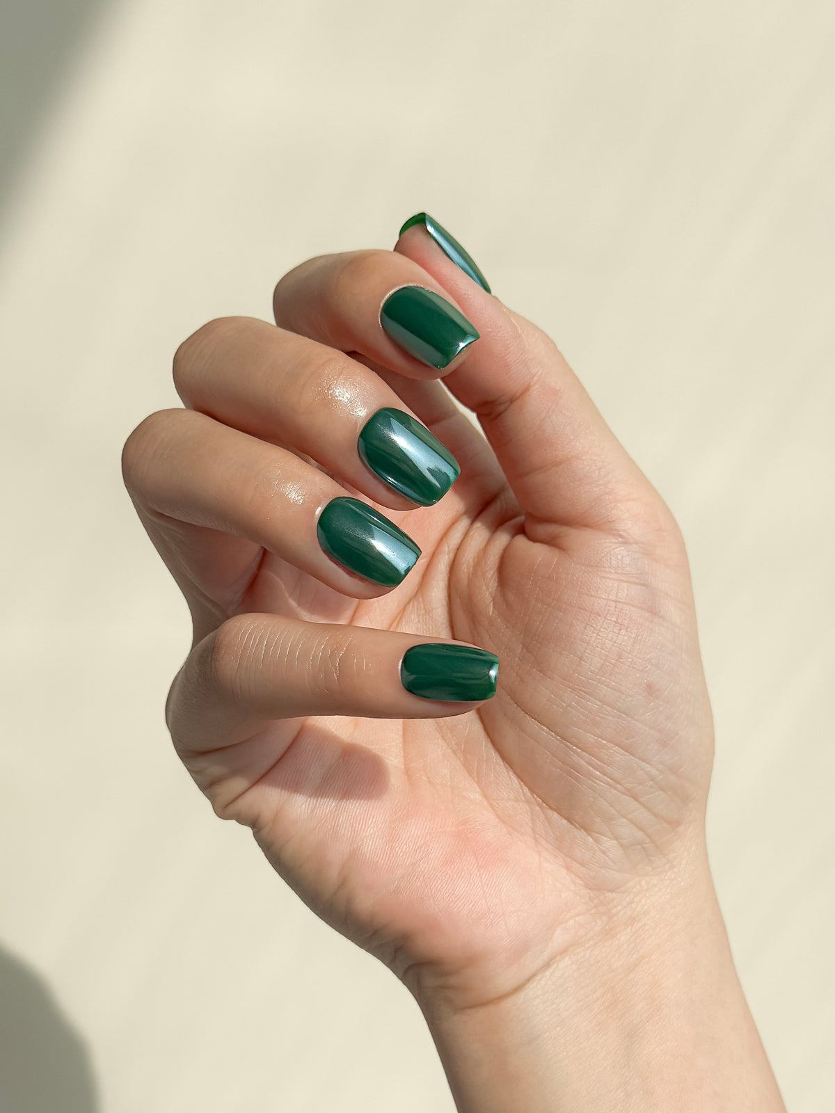 A hand wearing Promotion glossy dark forest green short square press-on nails in warm natural sunlight.