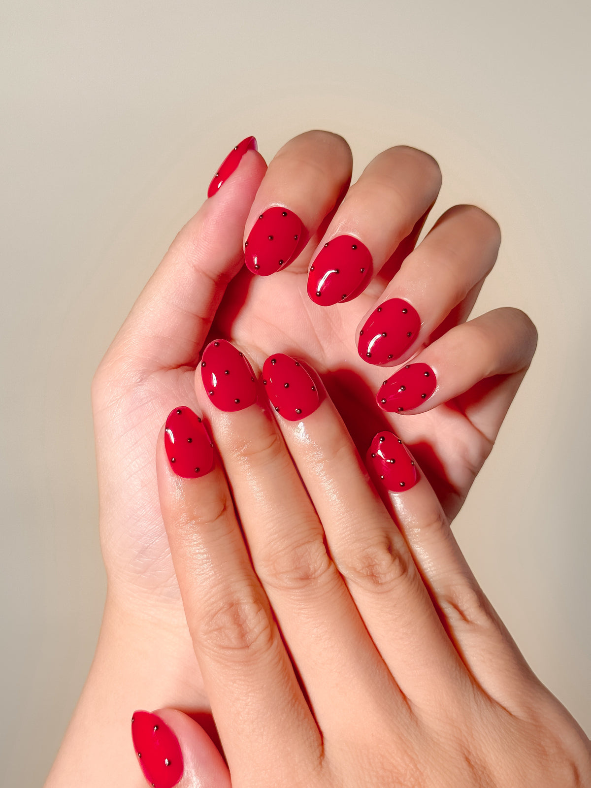Two hands wearing Strike One vivid red 3D stud extra-short oval press-on nails with fine silver lines.