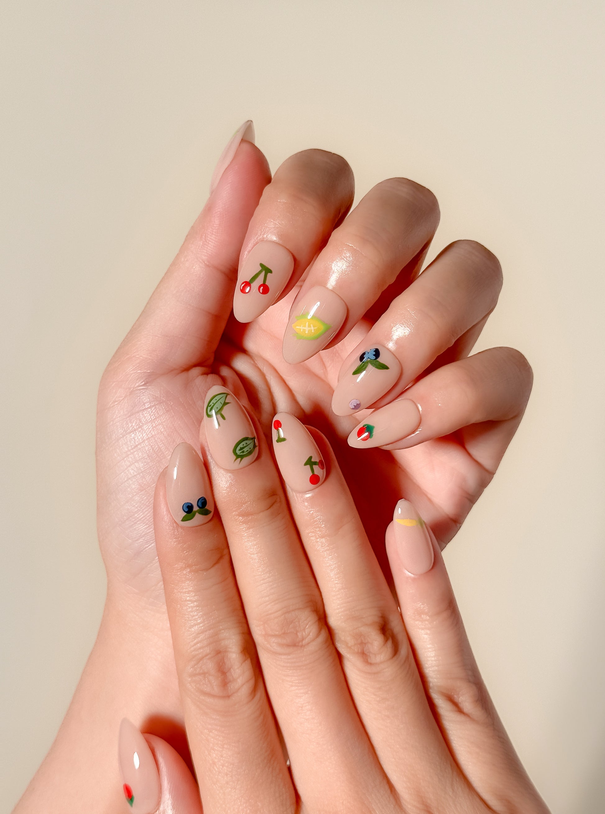 Summer Apron short almond press-on nails worn on two hands, featuring a glossy nude base with hand-painted cherries, blueberries, leafy vegetables, citrus, flowers, and colourful garden-bug illustrations.