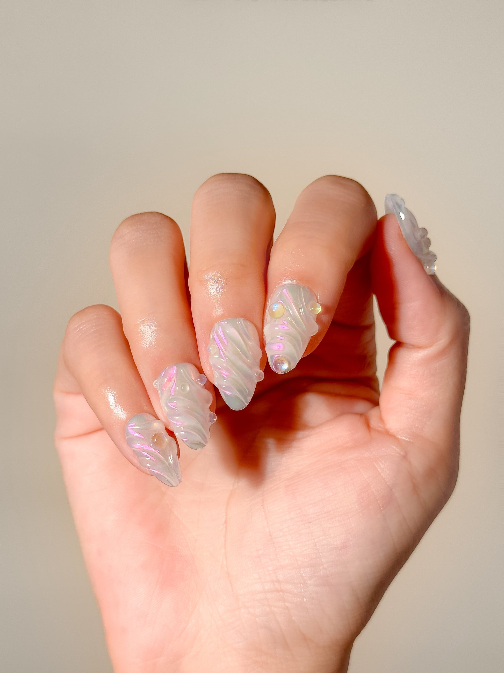 The Encore pearlescent short almond press-on nails worn on one hand, showing sculpted white swirls and pink-green iridescent shine.