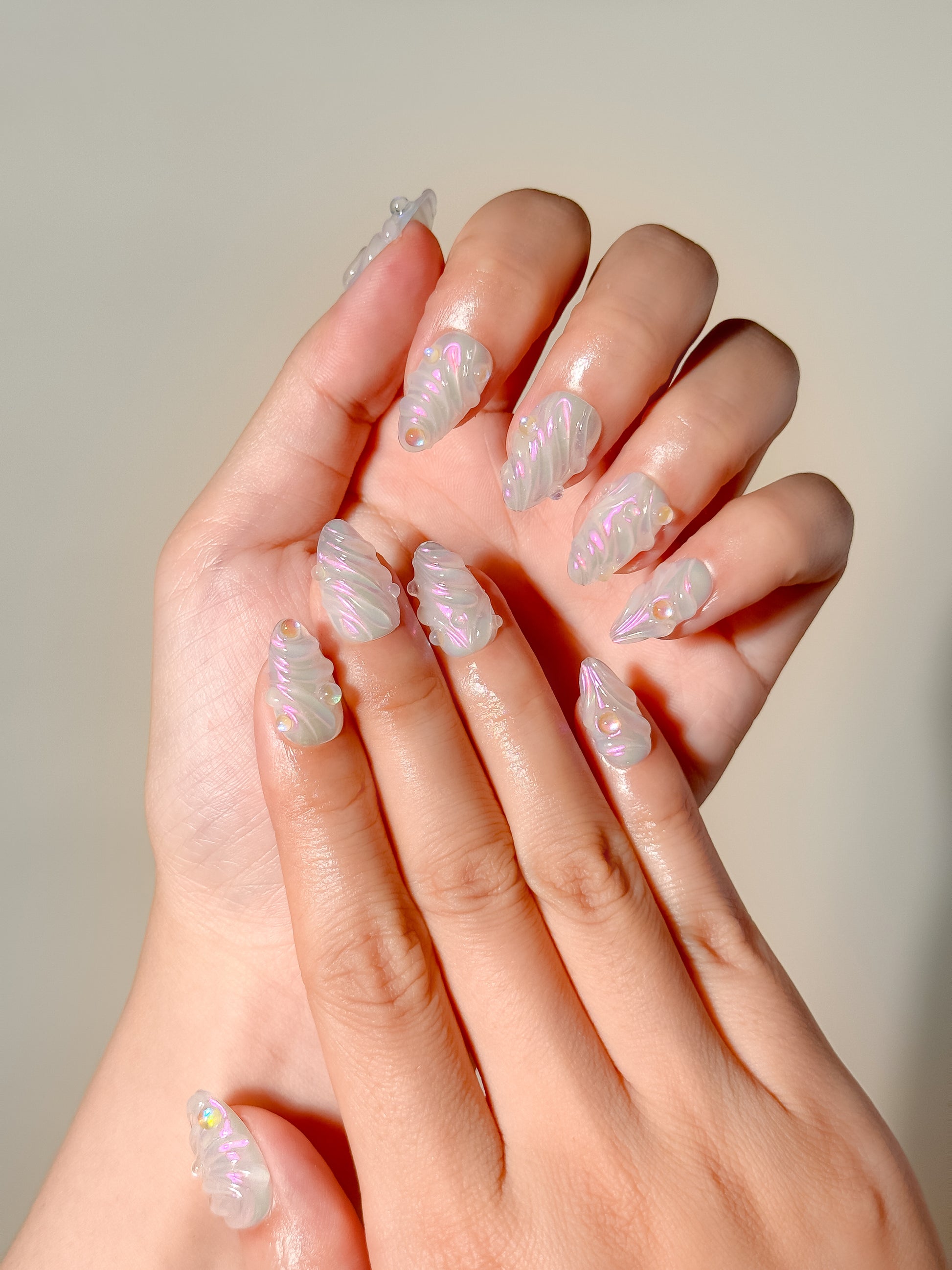 The Encore handmade 3D press-on nails worn on two hands, with translucent short almond tips, pearly swirls and gold accents.
