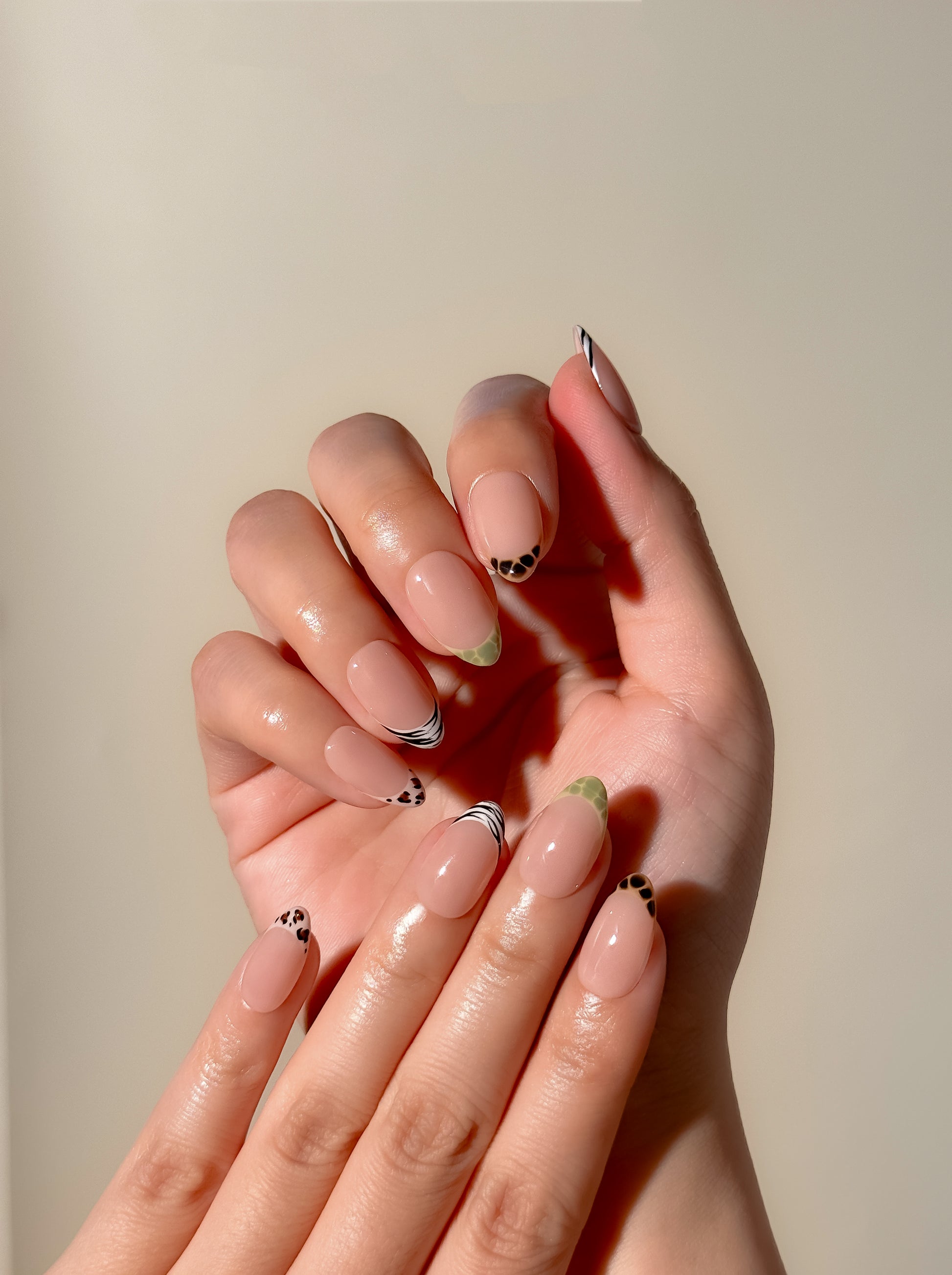 The Wild Card short almond press-on nails worn on two hands, featuring mixed animal-print French tips over a glossy sheer blush-pink base.