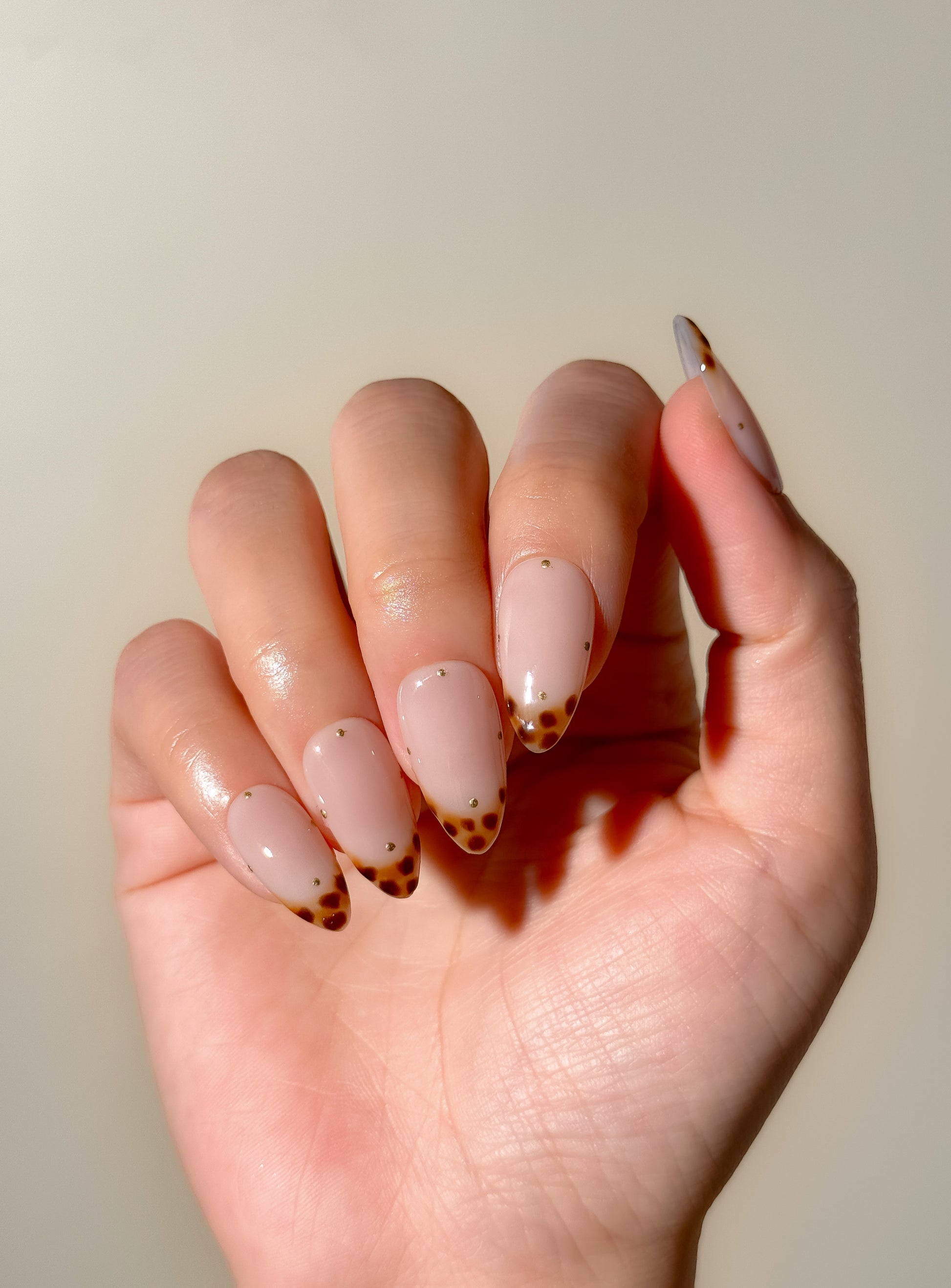 Tortoiseshell Tiny medium almond press-on nails worn on hand, featuring glossy tortoiseshell skinny French tips and scattered gold dots over a sheer blush-pink base.
