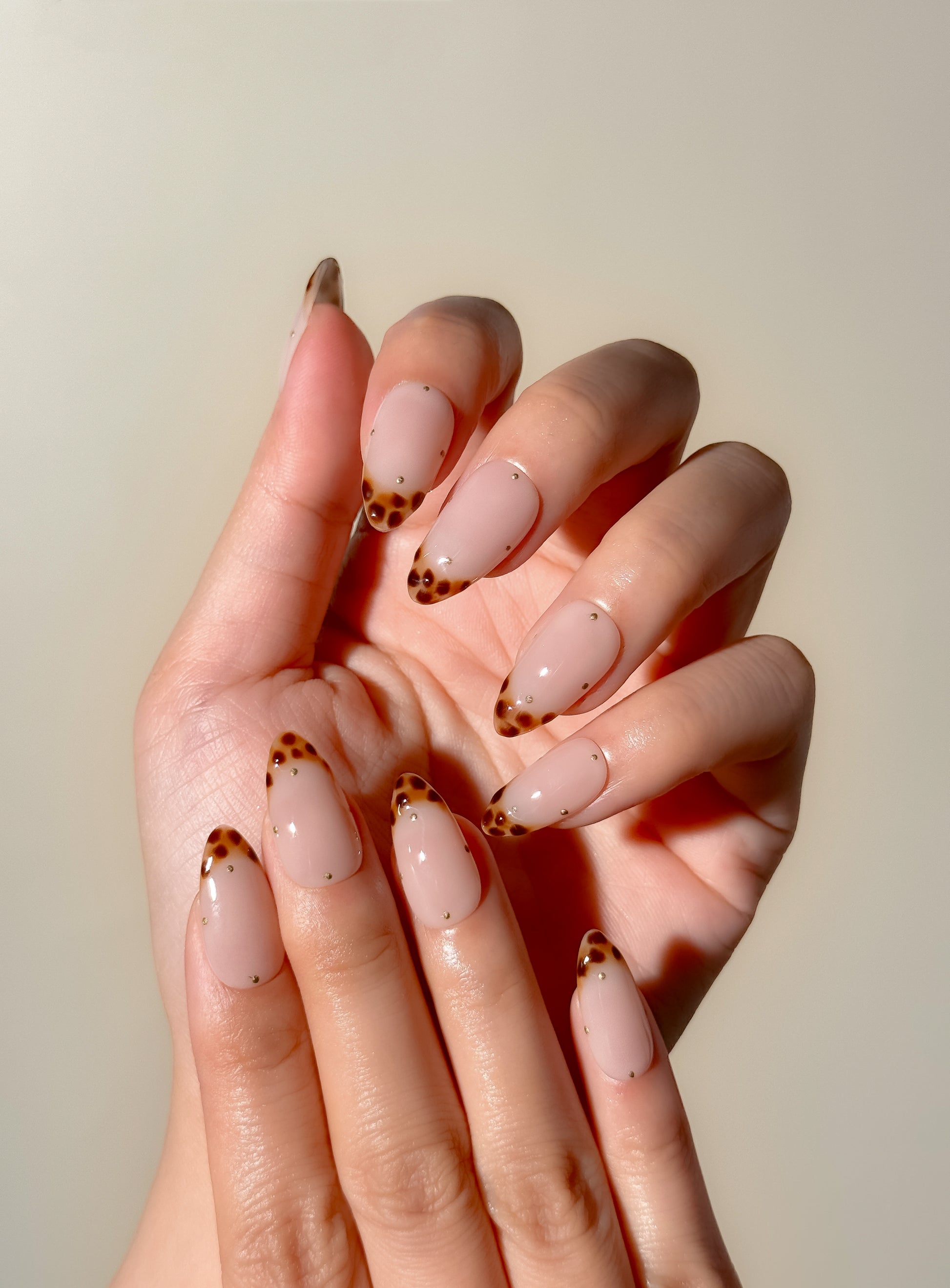 Tortoiseshell Tiny handmade press-on nails worn on two hands, featuring medium almond tortoiseshell French tips with delicate gold dot detailing.